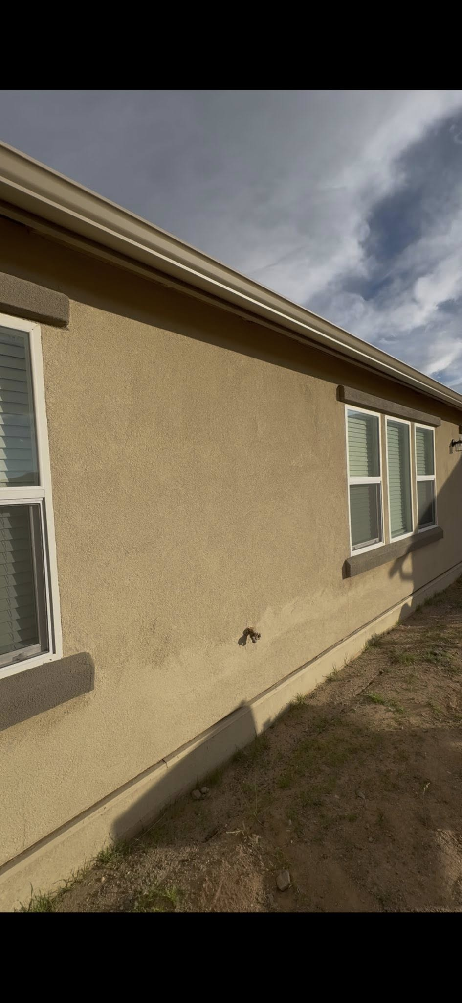 Exterior of a house with light brown stucco walls, windows, and a tan gutter under a cloudy sky.