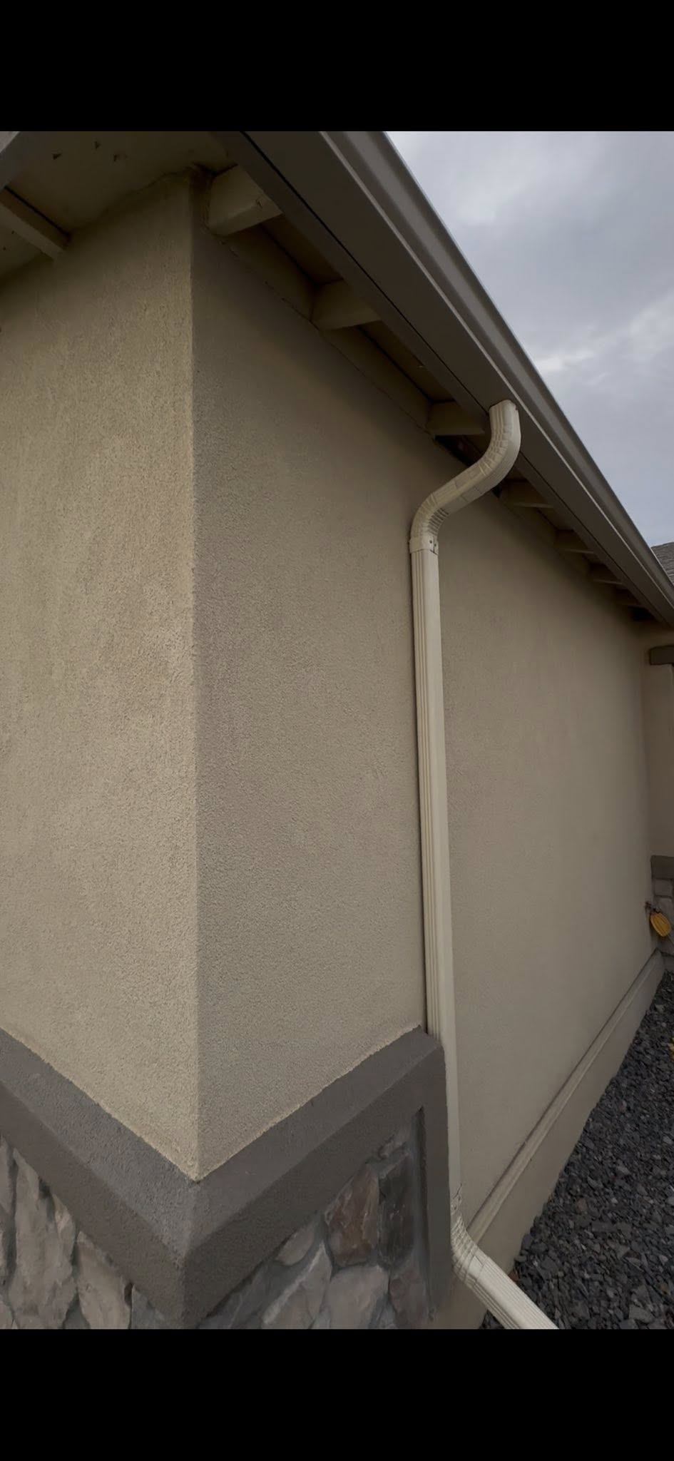 Exterior view of a stucco building with a downspout and gutter against a cloudy sky.