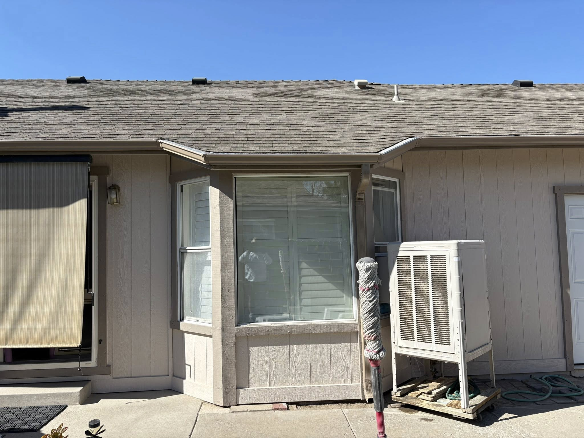 Exterior view of a building with a window, awning, and a swamp cooler. Tan siding and a brown roof.