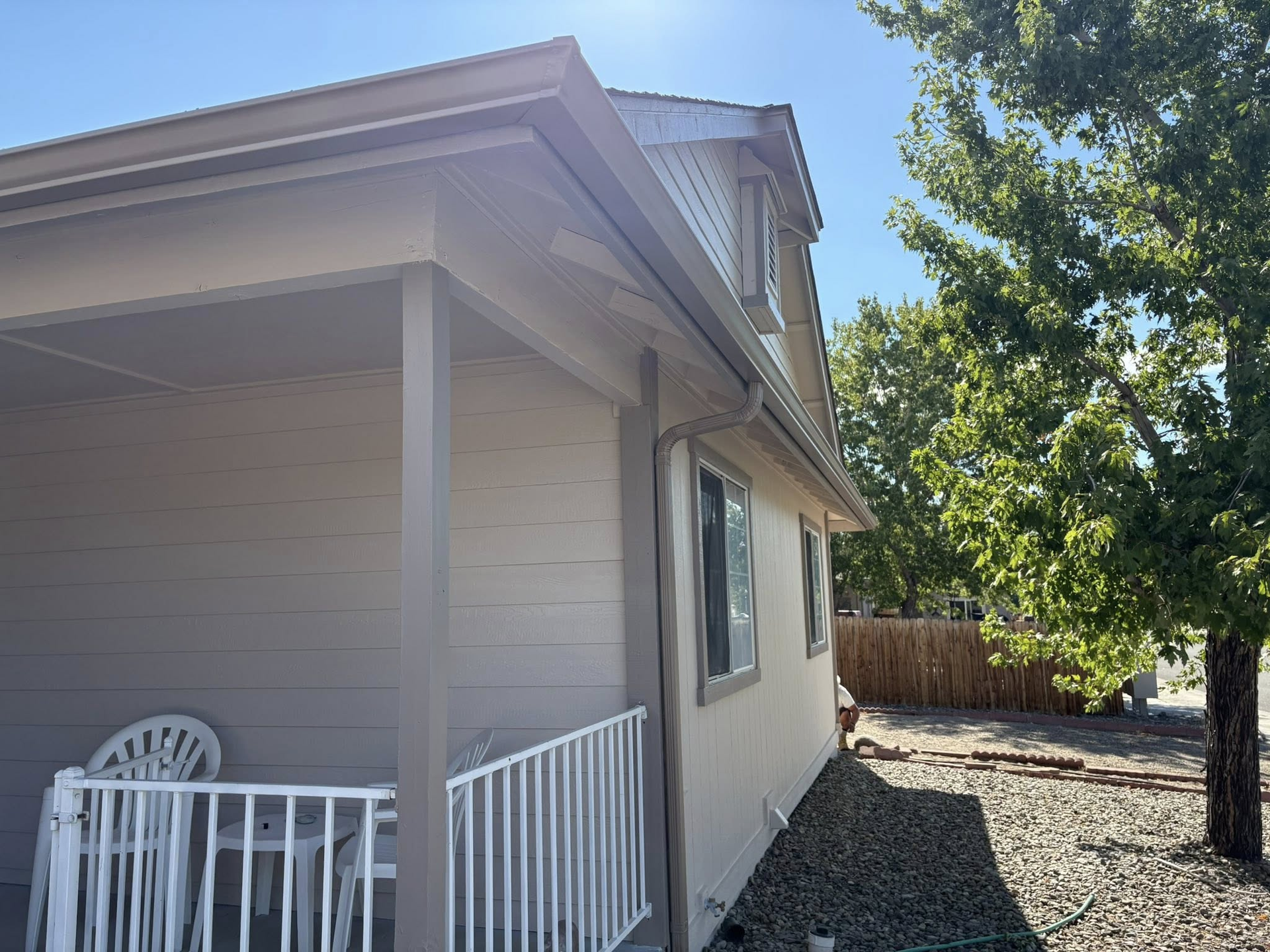 Beige house with a porch and white railing, under a blue sky, and a tree to the right.