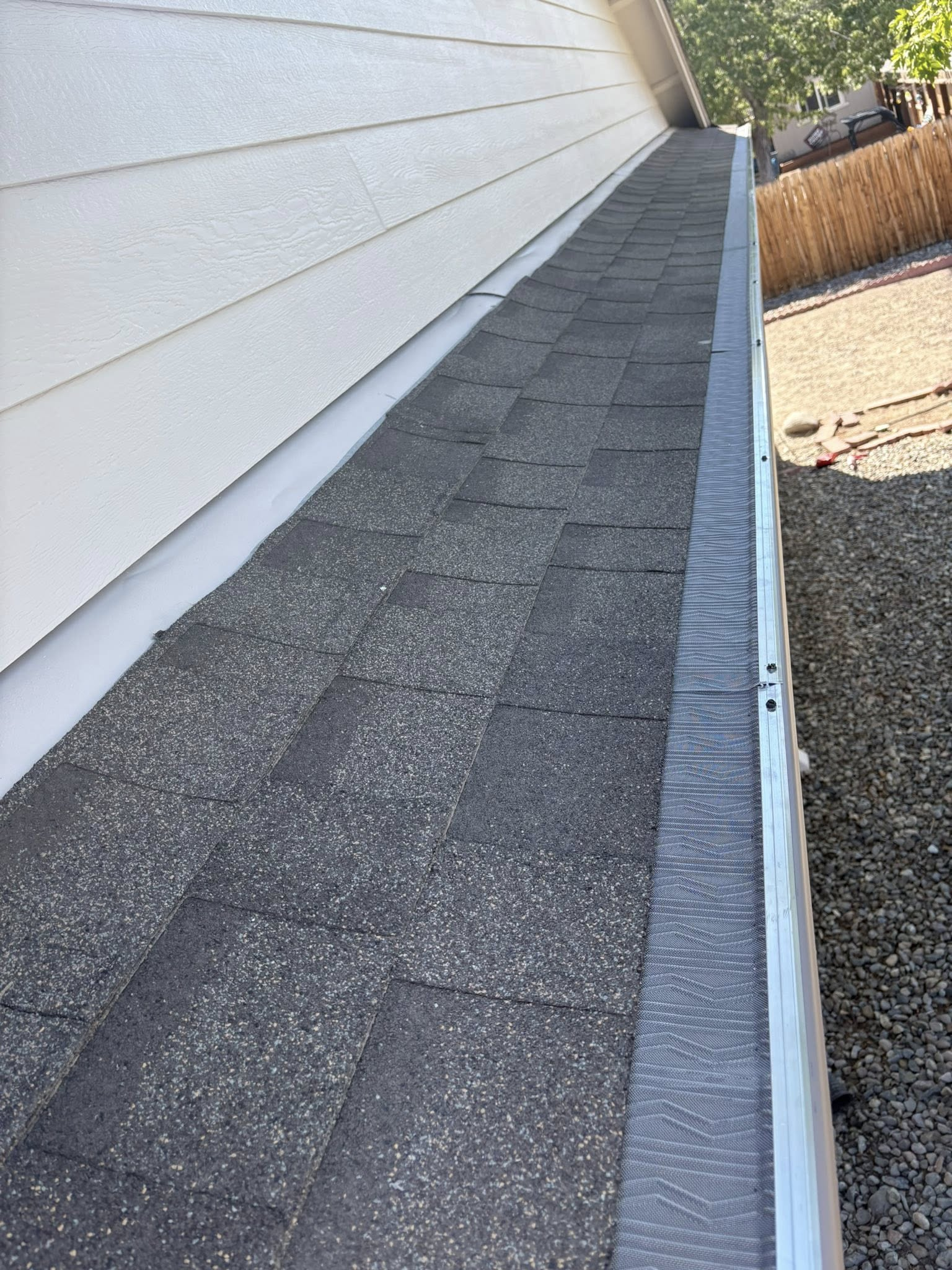 Close-up of a roof edge with gray shingles, white trim, and a gutter, under a blue sky.