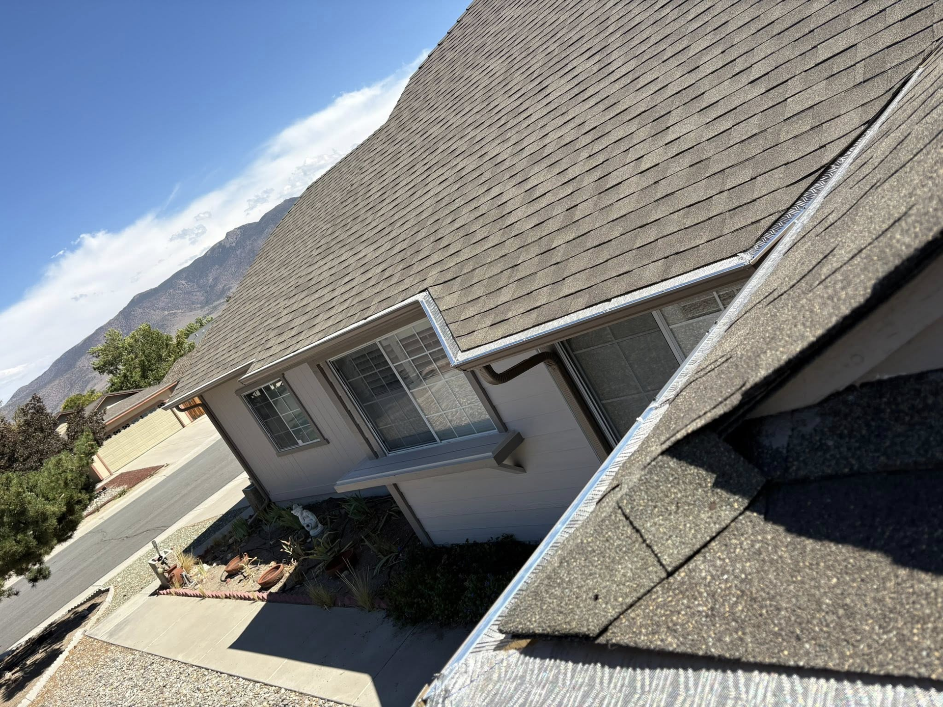 View of a house with a gray shingle roof, siding, and nearby mountain under a blue sky.