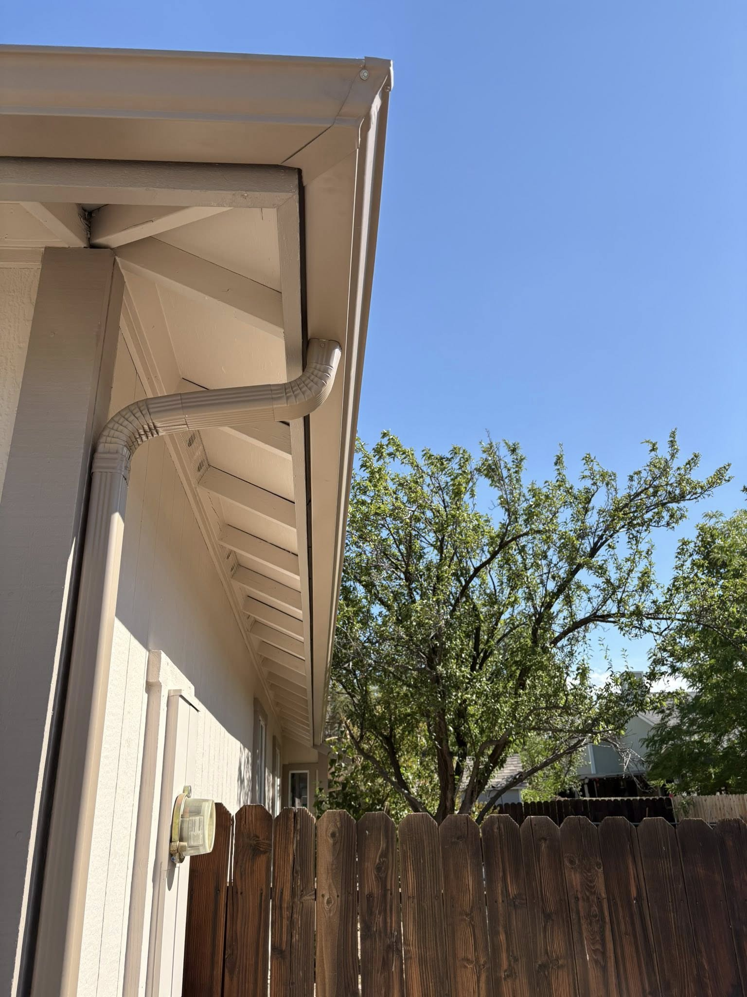 Corner of a beige house with a gutter, white soffit, and wooden fence, with a tree and blue sky in the background.
