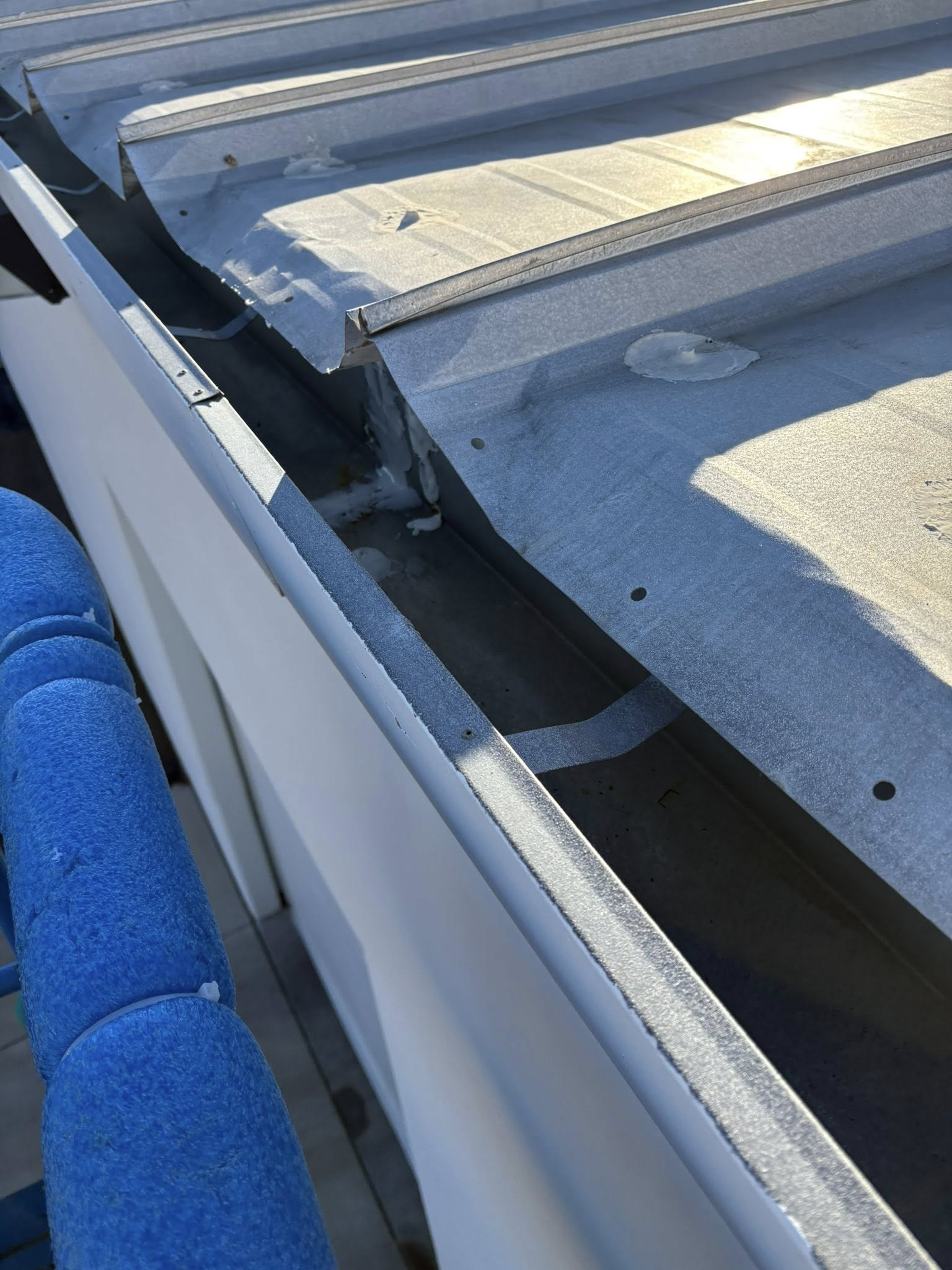 Gutter on a metal roof, close up.  White gutter next to the silver roof; blue, textured column is on left.