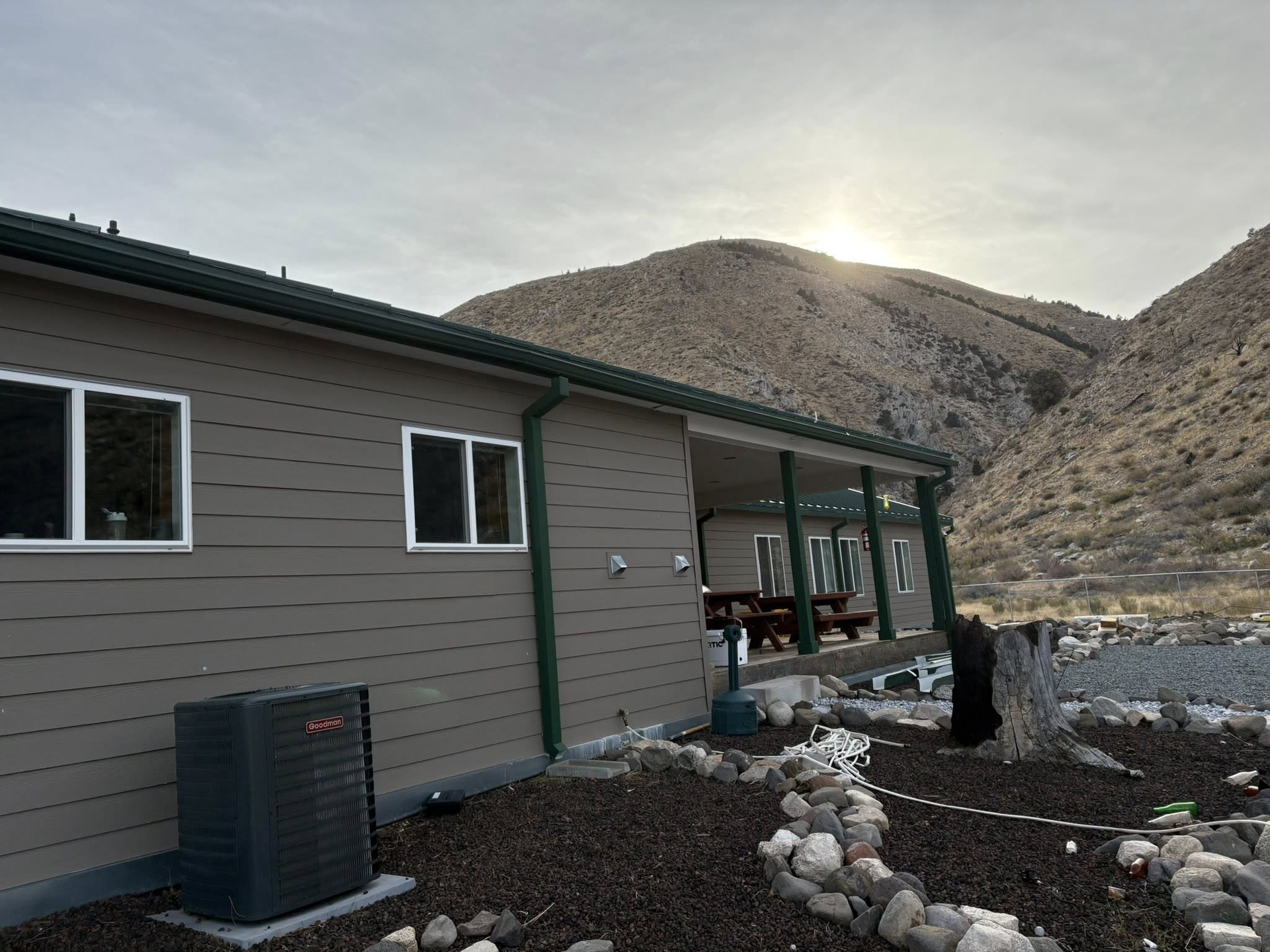A tan house with a green trim and covered porch is set against a backdrop of mountains under a cloudy sky.