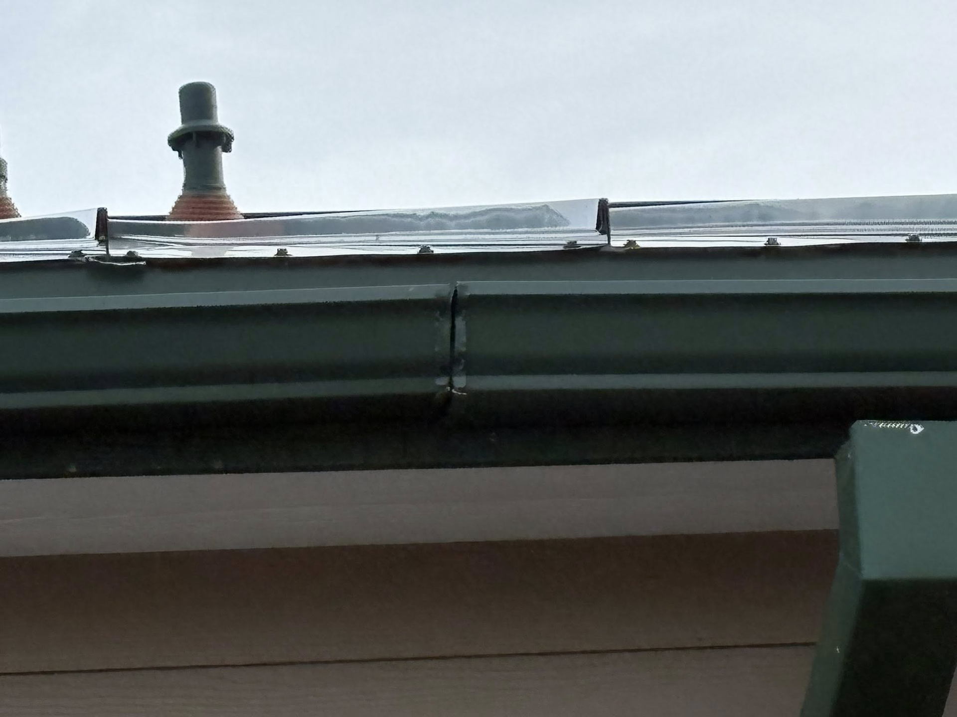Green rain gutter on a light-colored building with dark roof and chimney vents.