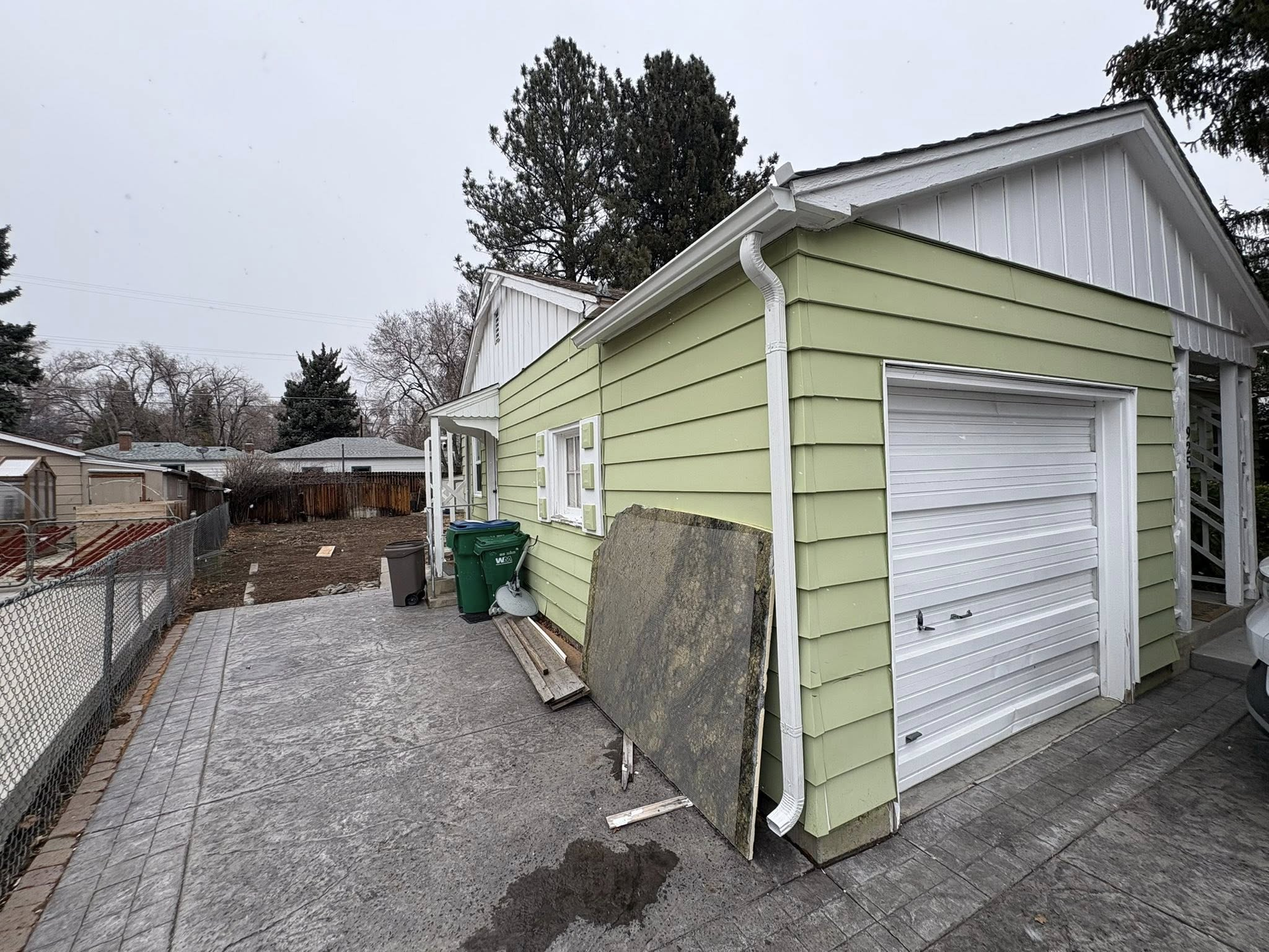 Green-sided house with white garage door. Driveway with debris. Cloudy sky.