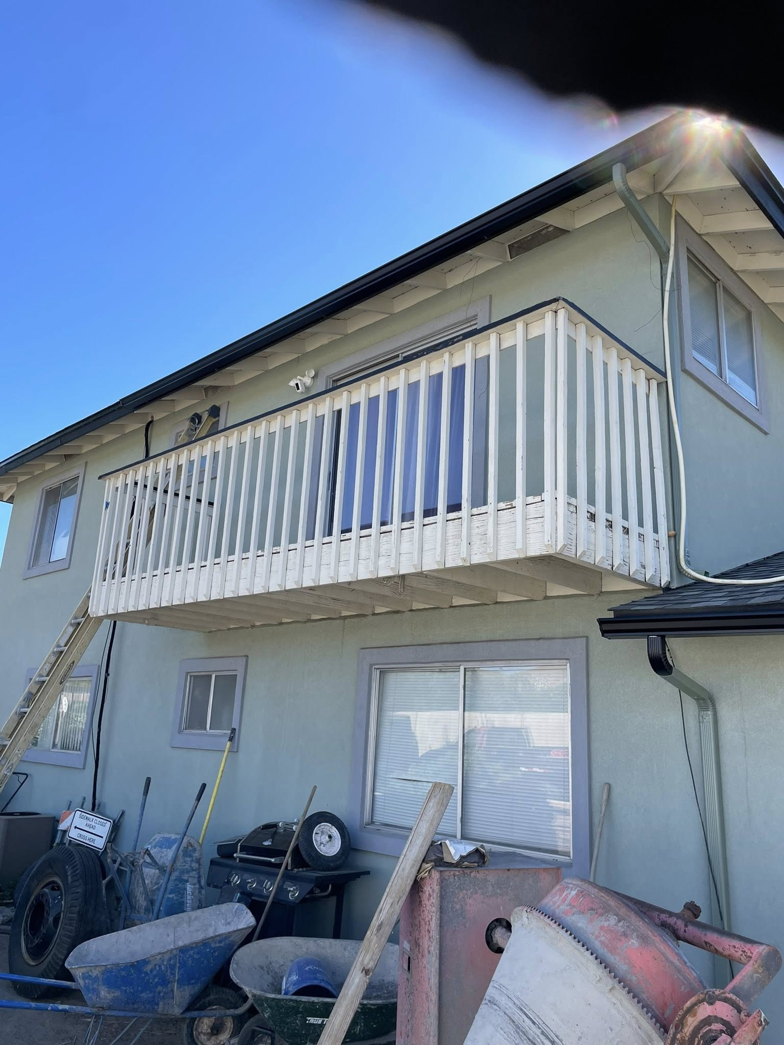 Two-story house with white balcony and railing. Construction materials and tools are visible below.