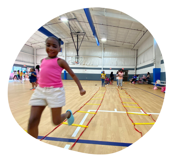 Girl runs agility ladder in gymnasium. She wears pink tank top, white shorts, smiling. Others watching.