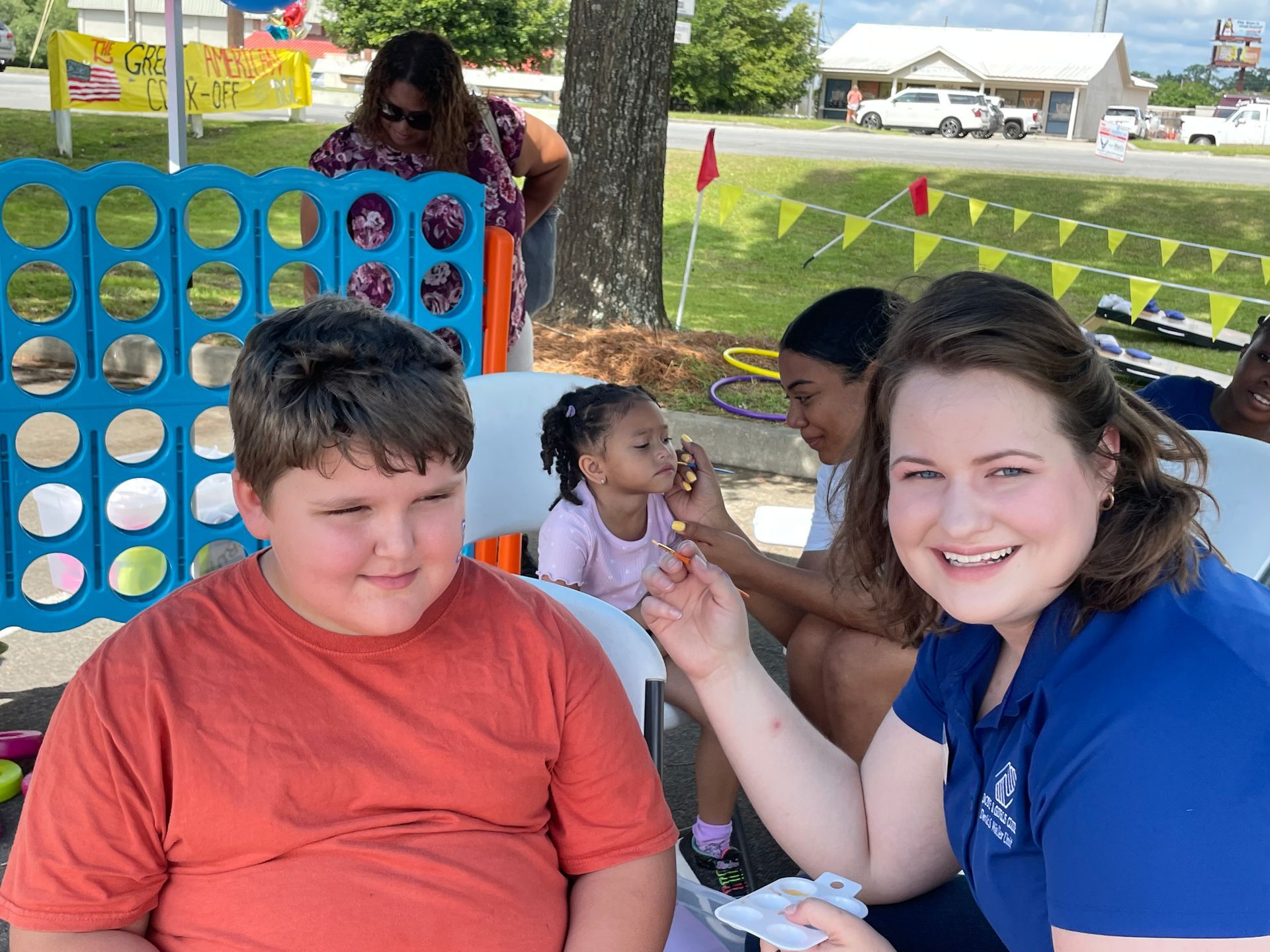 Boy and woman smiling. A girl has face paint applied at an outdoor event with games.