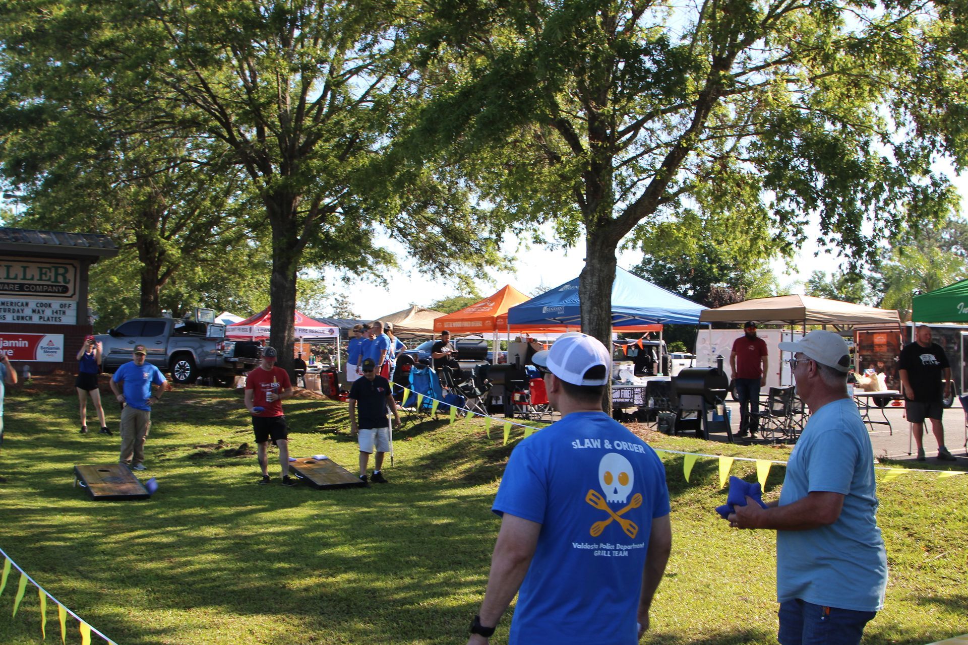 People playing cornhole in a park, with tents and vendors in the background on a sunny day.