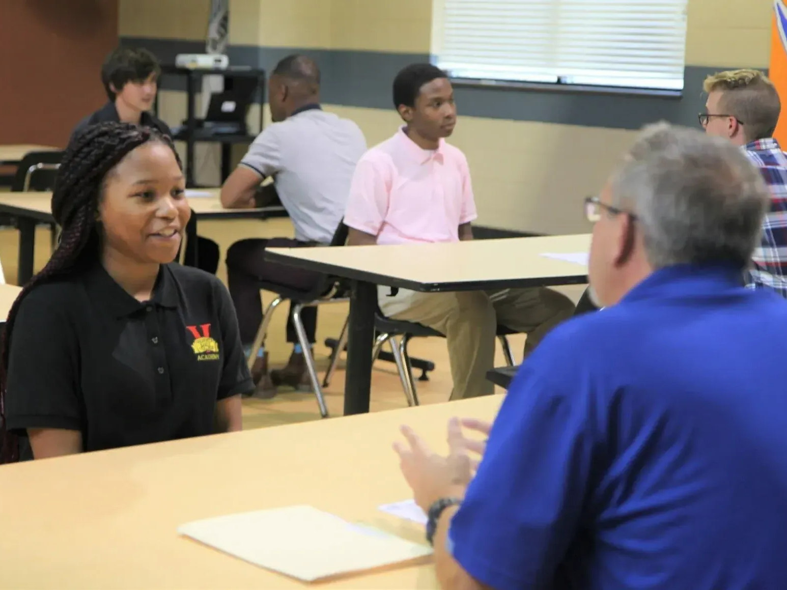 Student in a black shirt is being interviewed; people sit at tables in a classroom-like setting.