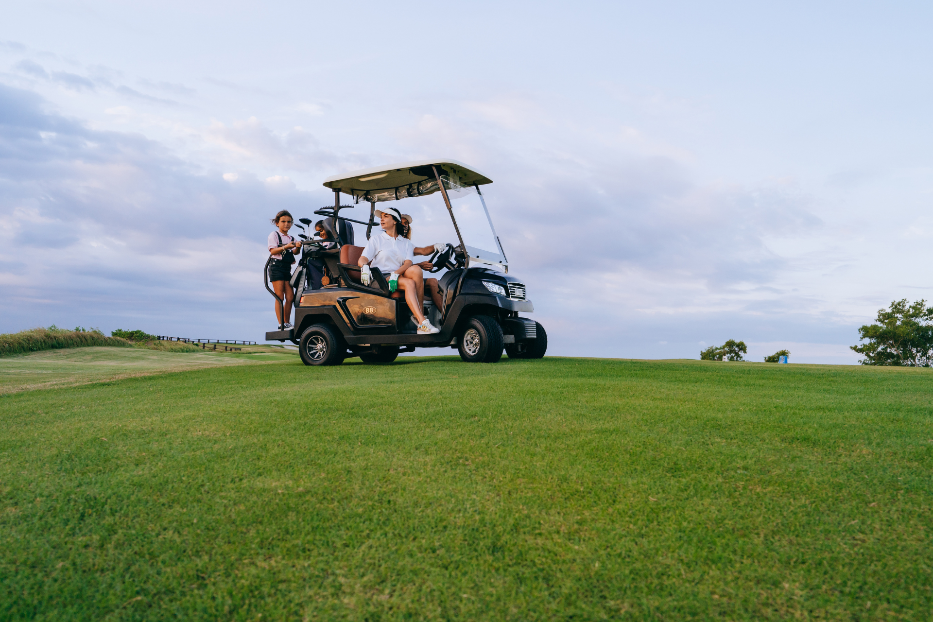Golfers in a golf cart on a green course. Two women are on the cart, another is on the back.