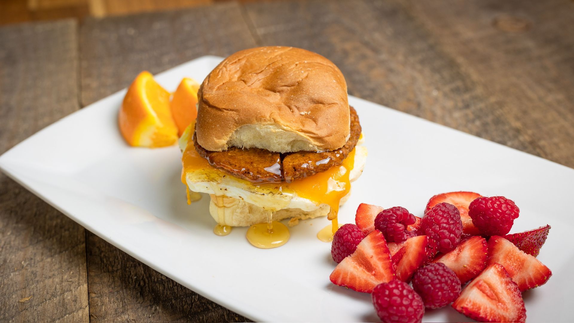 Breakfast sandwich with fruit on a white plate: egg, cheese, sausage, bun, strawberries, raspberries, orange slices.