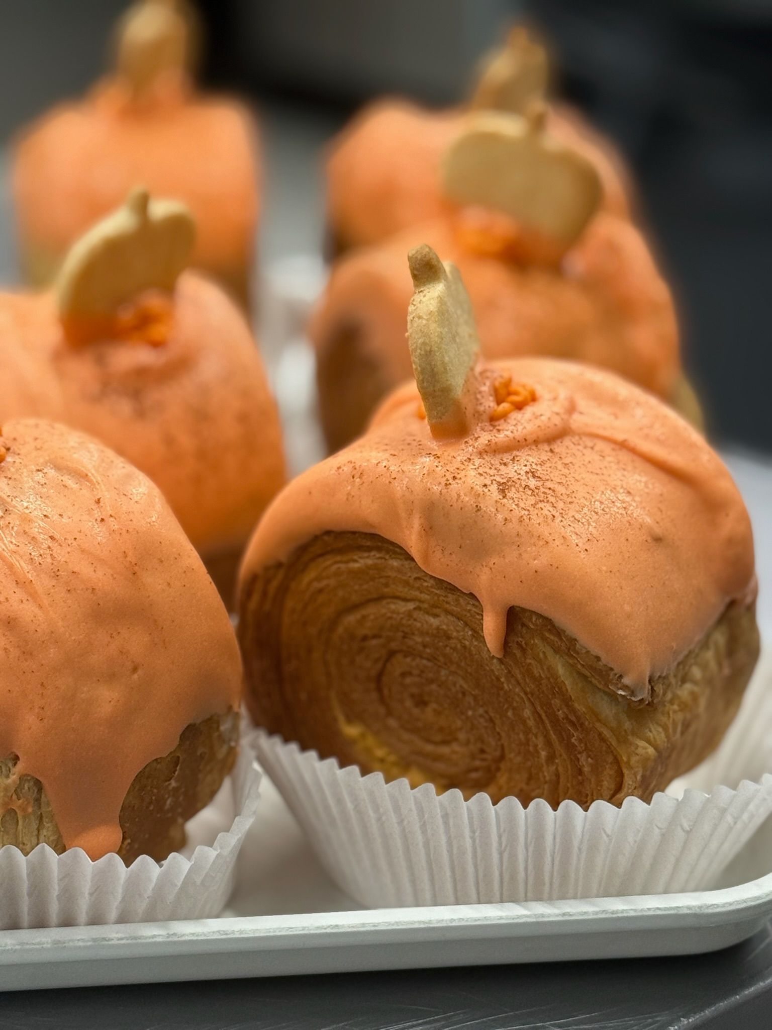 Pumpkin-shaped pastries with orange frosting, topped with a biscuit stem. In white paper liners, on a tray.
