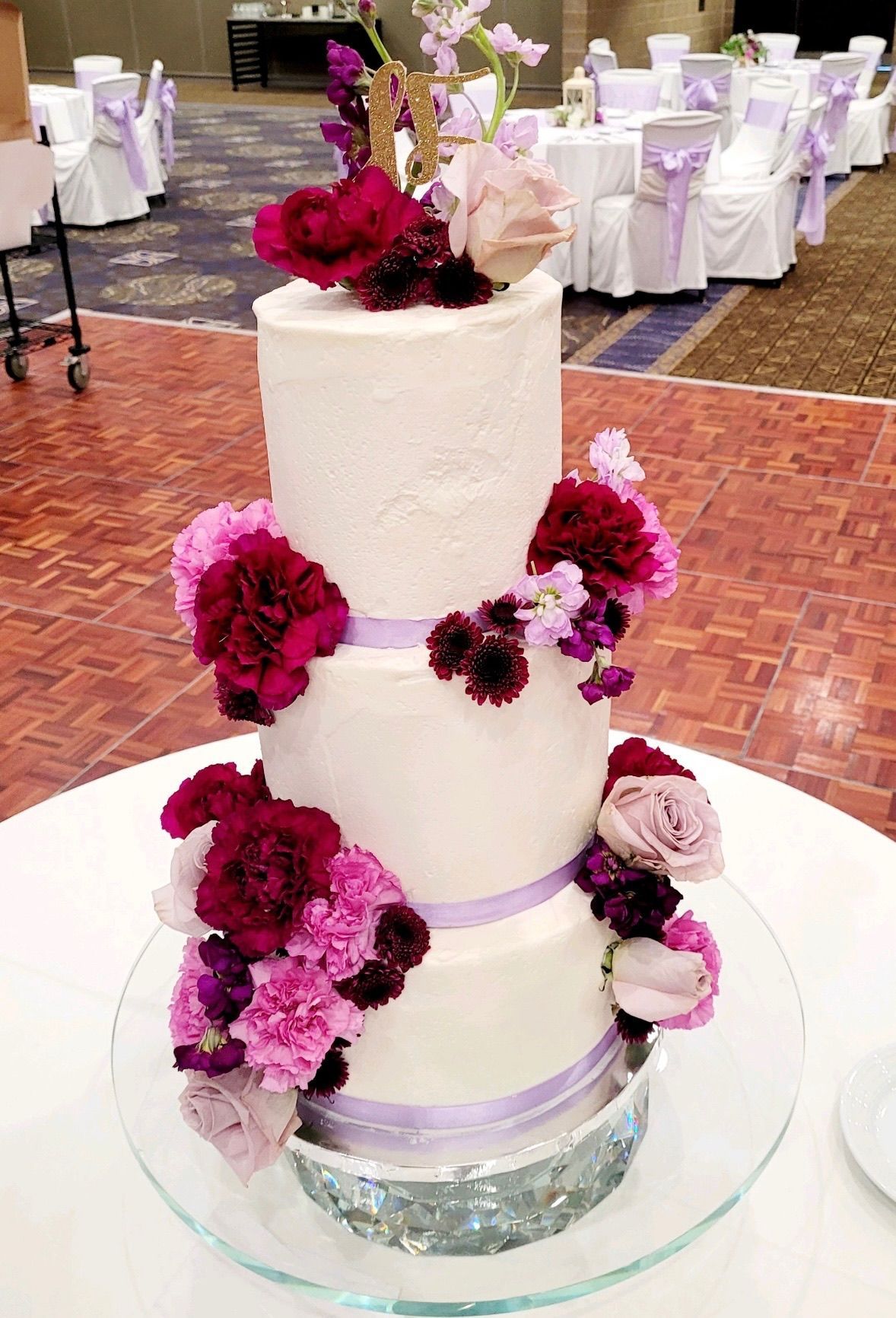Three-tiered wedding cake, decorated with flowers and ribbons. Purple, pink, and burgundy flowers adorn the white frosted cake.