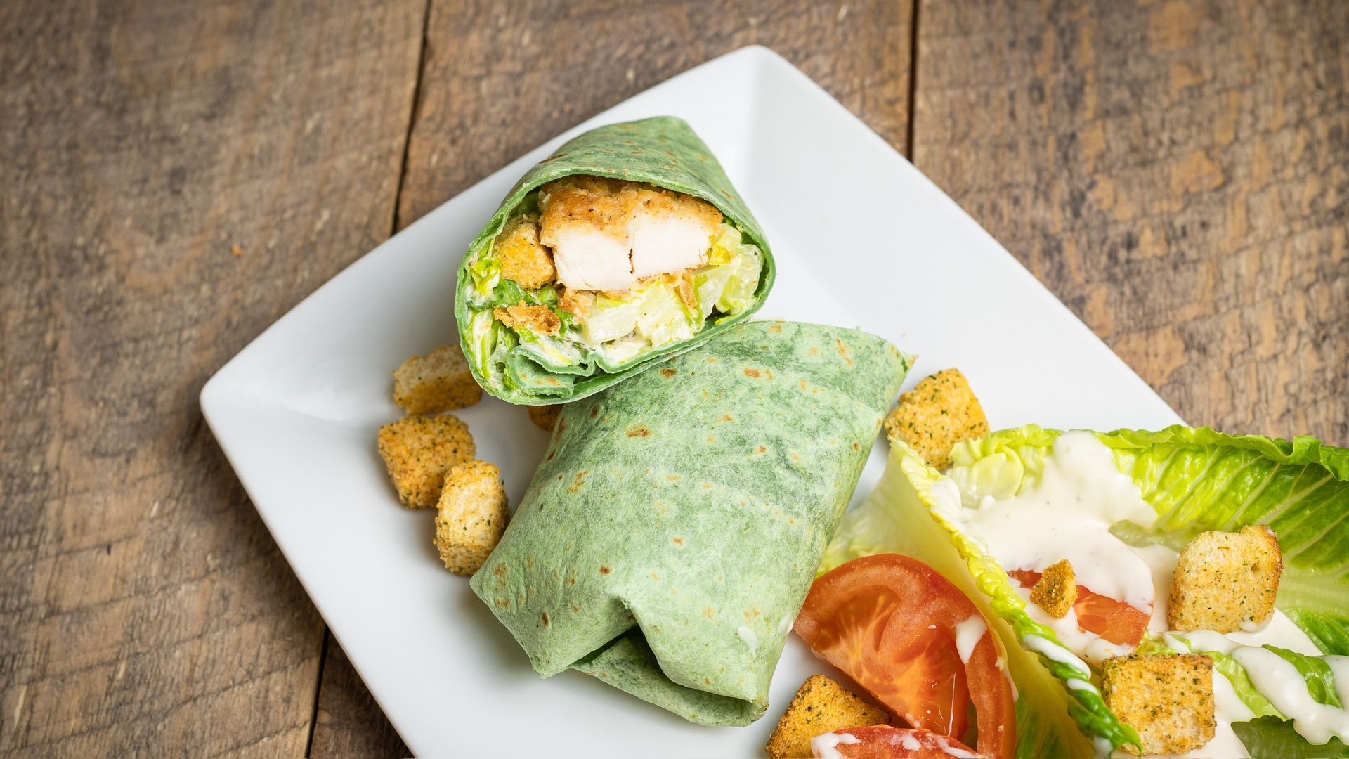 Green wrap sandwich with chicken and Caesar salad on a white plate.