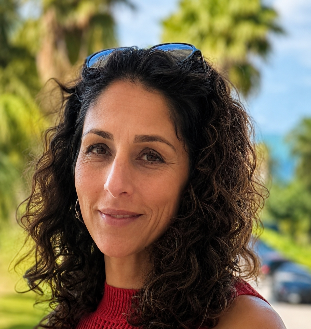 Woman with curly dark hair in a red top outdoors, with palm trees and blue sky behind her