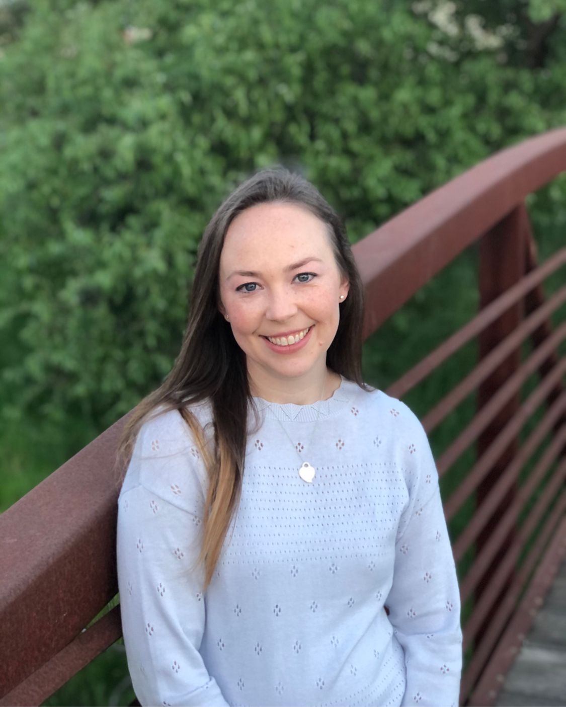Smiling person in a light blue sweater standing on a bridge with greenery behind them