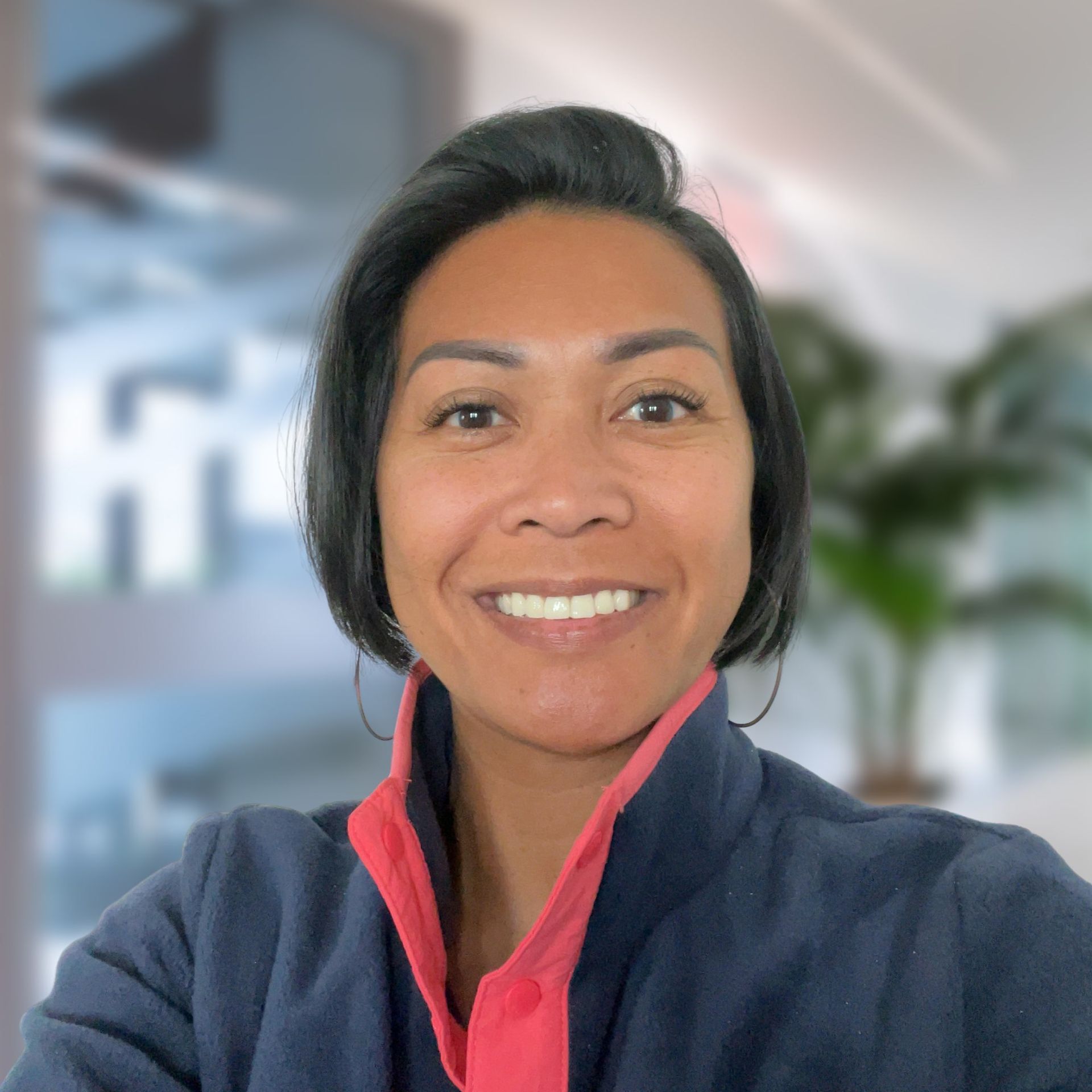 Smiling person in a navy jacket with a pink collar, in a blurred indoor office setting.