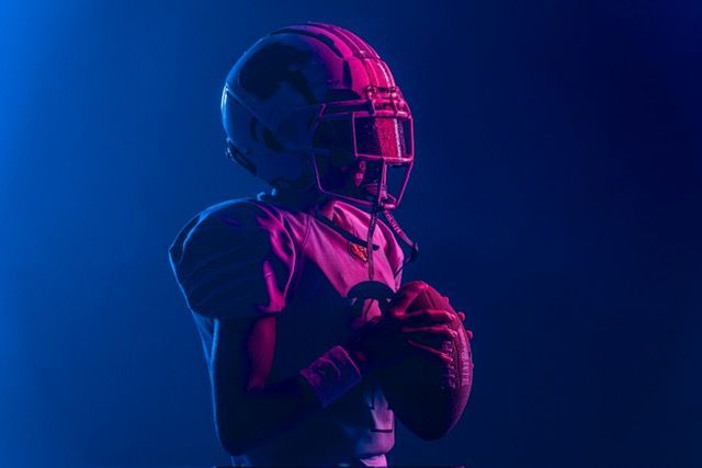 Football players huddle around a football, reaching hands together, blue sky backdrop.