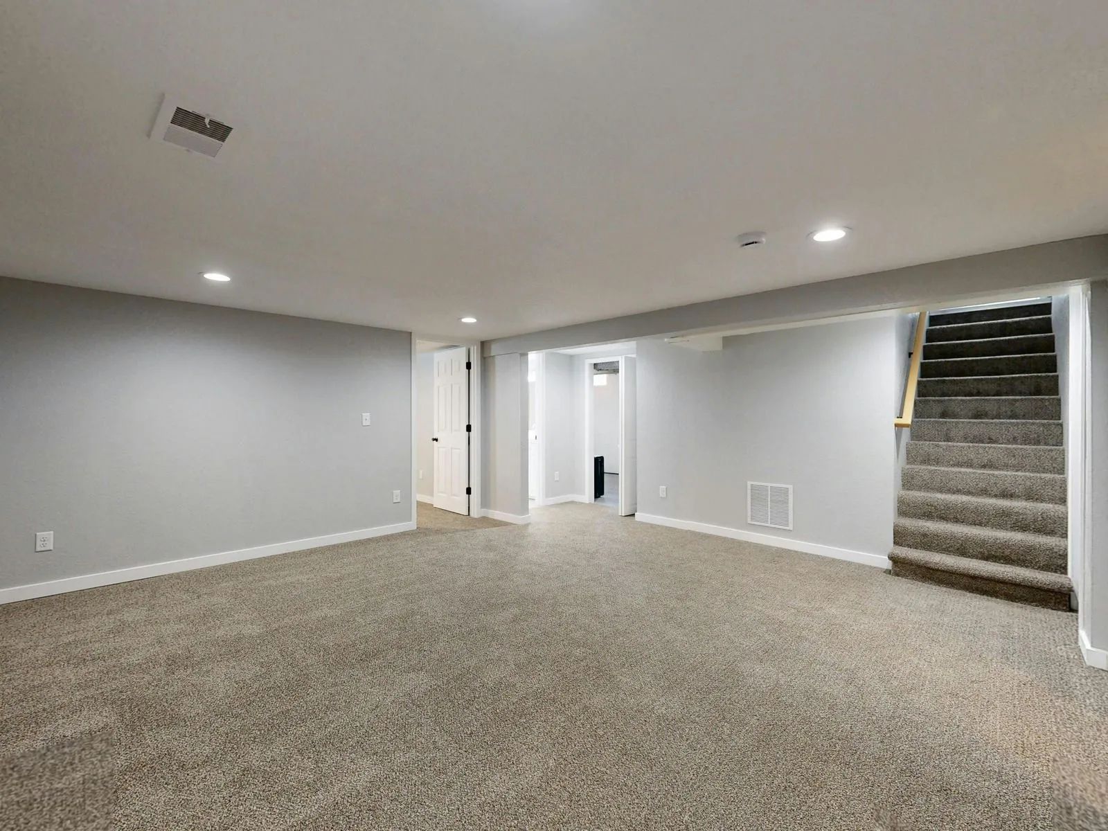 Empty carpeted basement room with gray walls, recessed lights, and stairs on the right