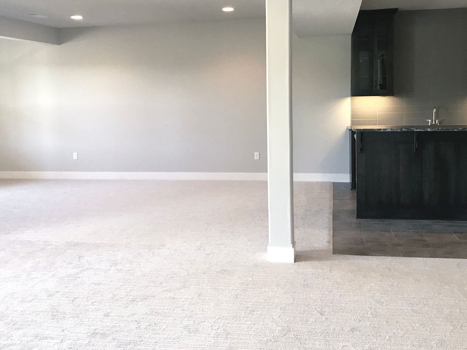 Empty beige living room with carpet and a partial view of a dark kitchen area