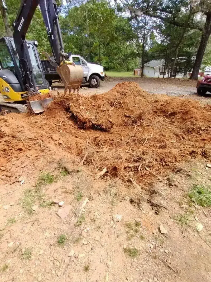 Excavator moving a large pile of brown mulch/dirt near a white pickup in a wooded area.