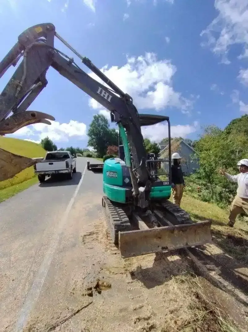 Excavator grading a roadside ditch beside traffic on a sunny day