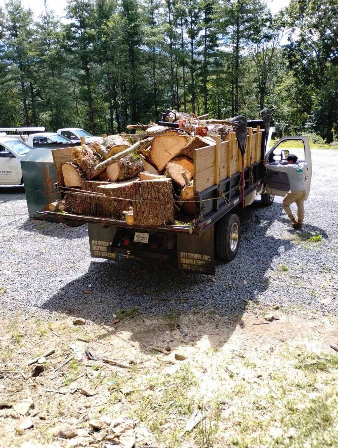 Small truck loaded with split firewood at a gravel pull-off, with a person beside it and trees in the background