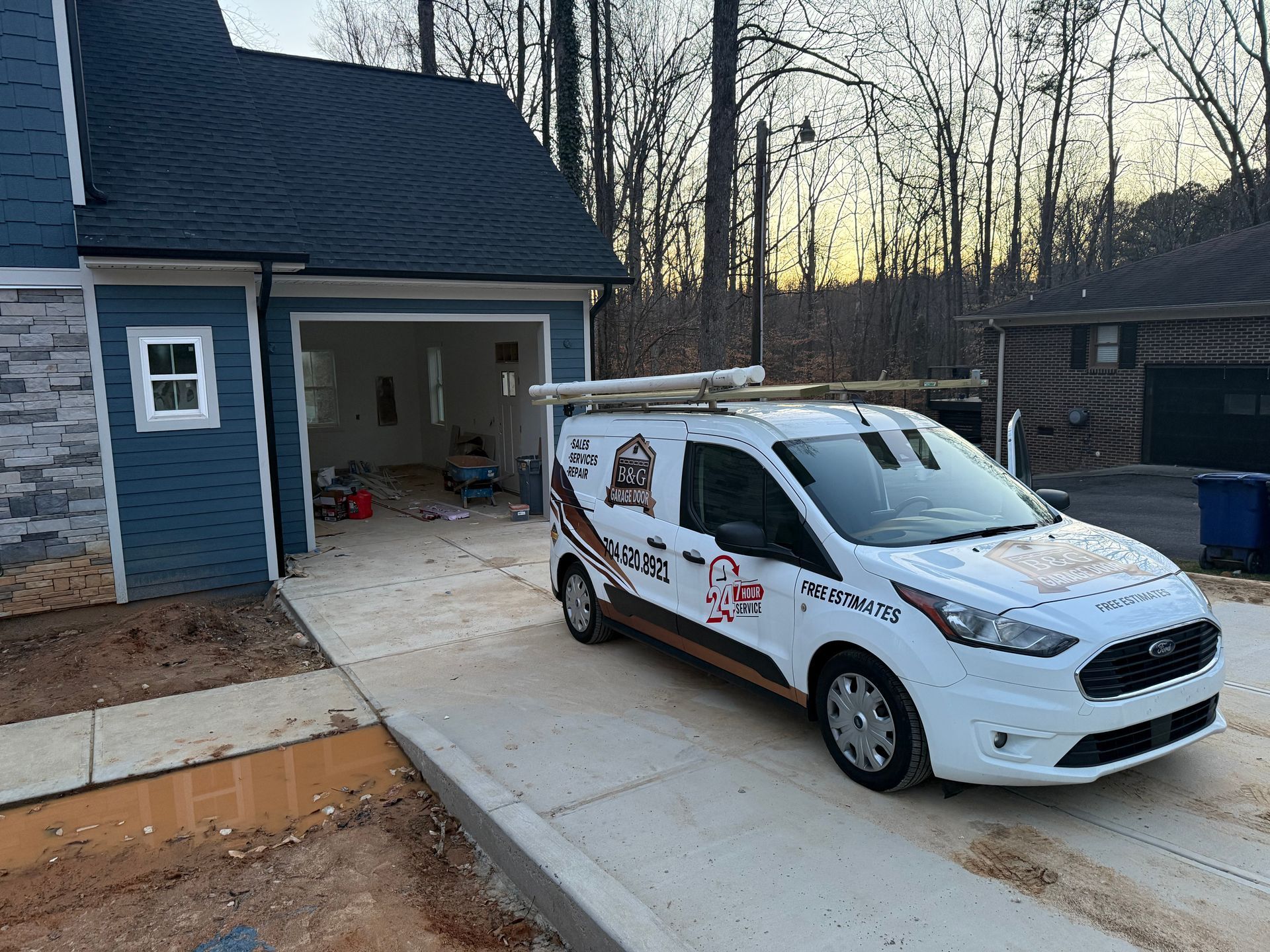 White service van parked in a driveway beside a blue house at dusk, garage door open.