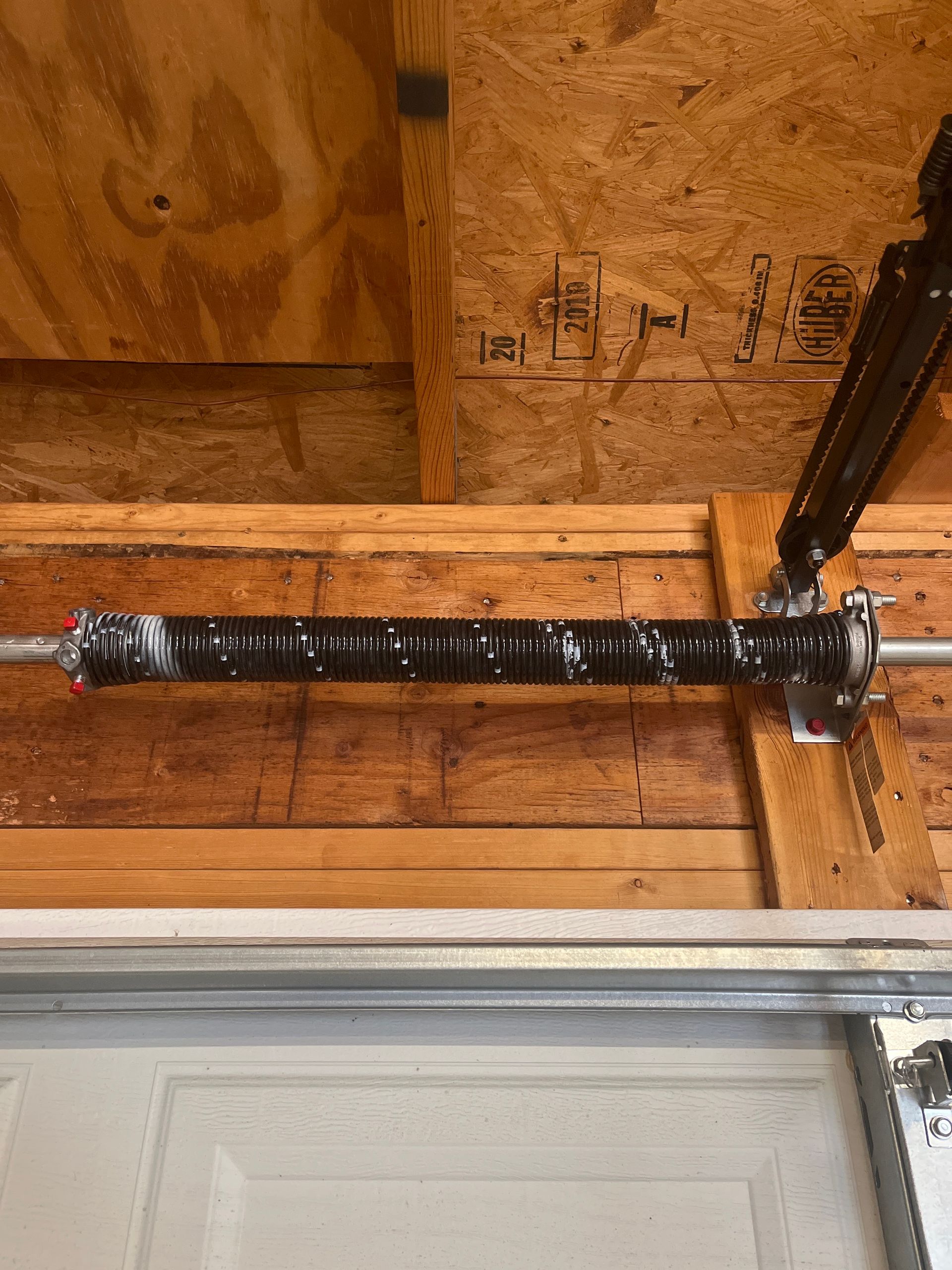 Black garage door torsion spring mounted horizontally on wooden framing in an unfinished attic space