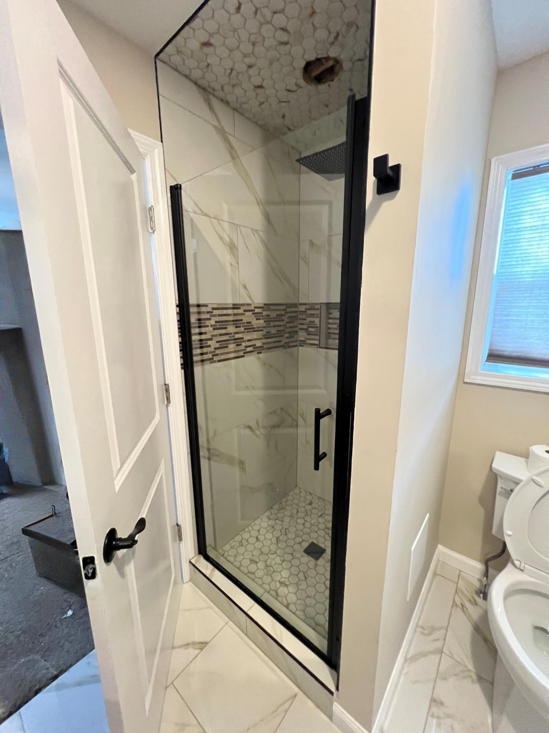 Bathroom with a glass shower stall, beige tile, and a white toilet beside a window.