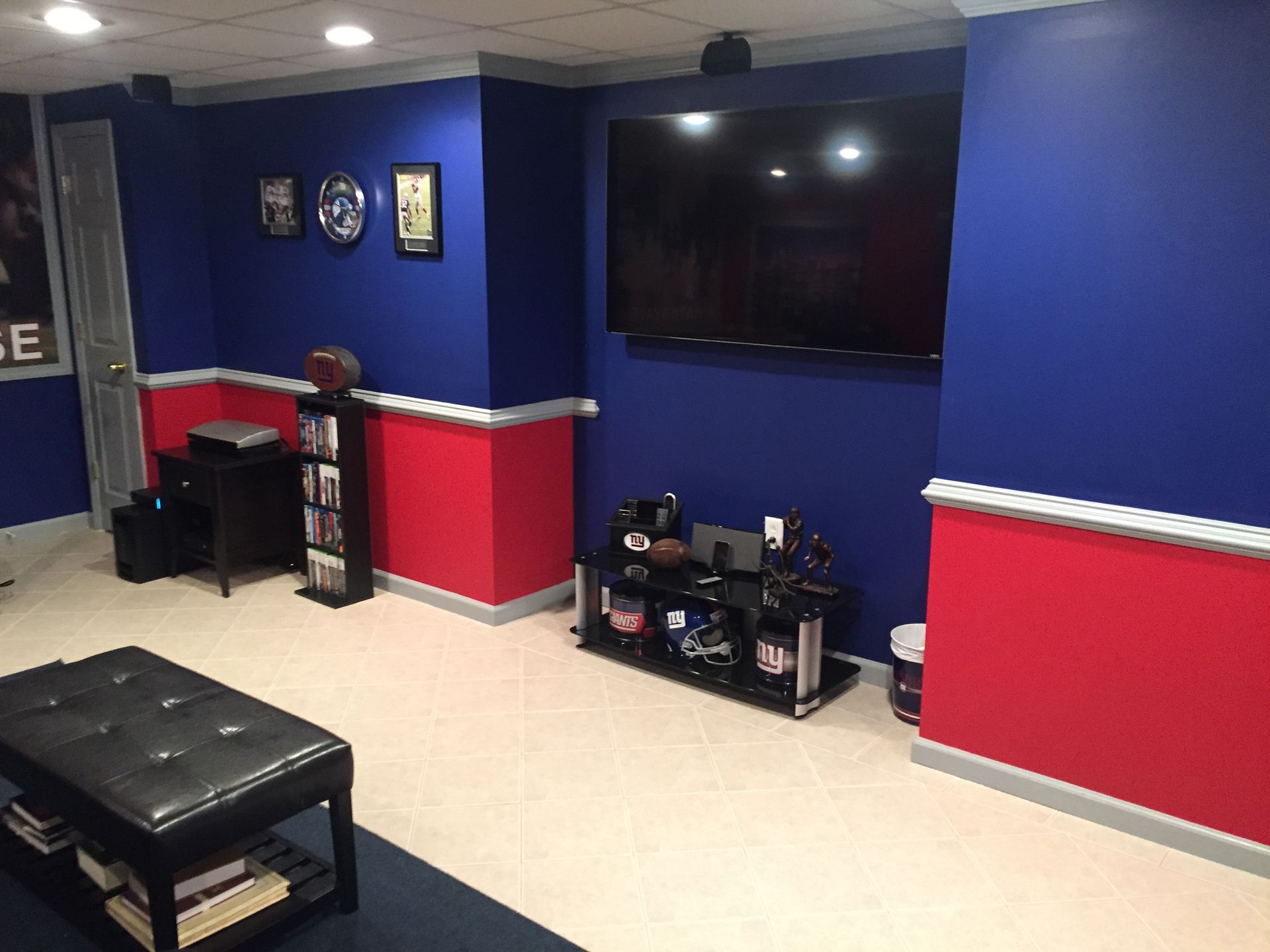 Basement game room with blue and red walls, wall-mounted TV, small desk, and black ottoman table