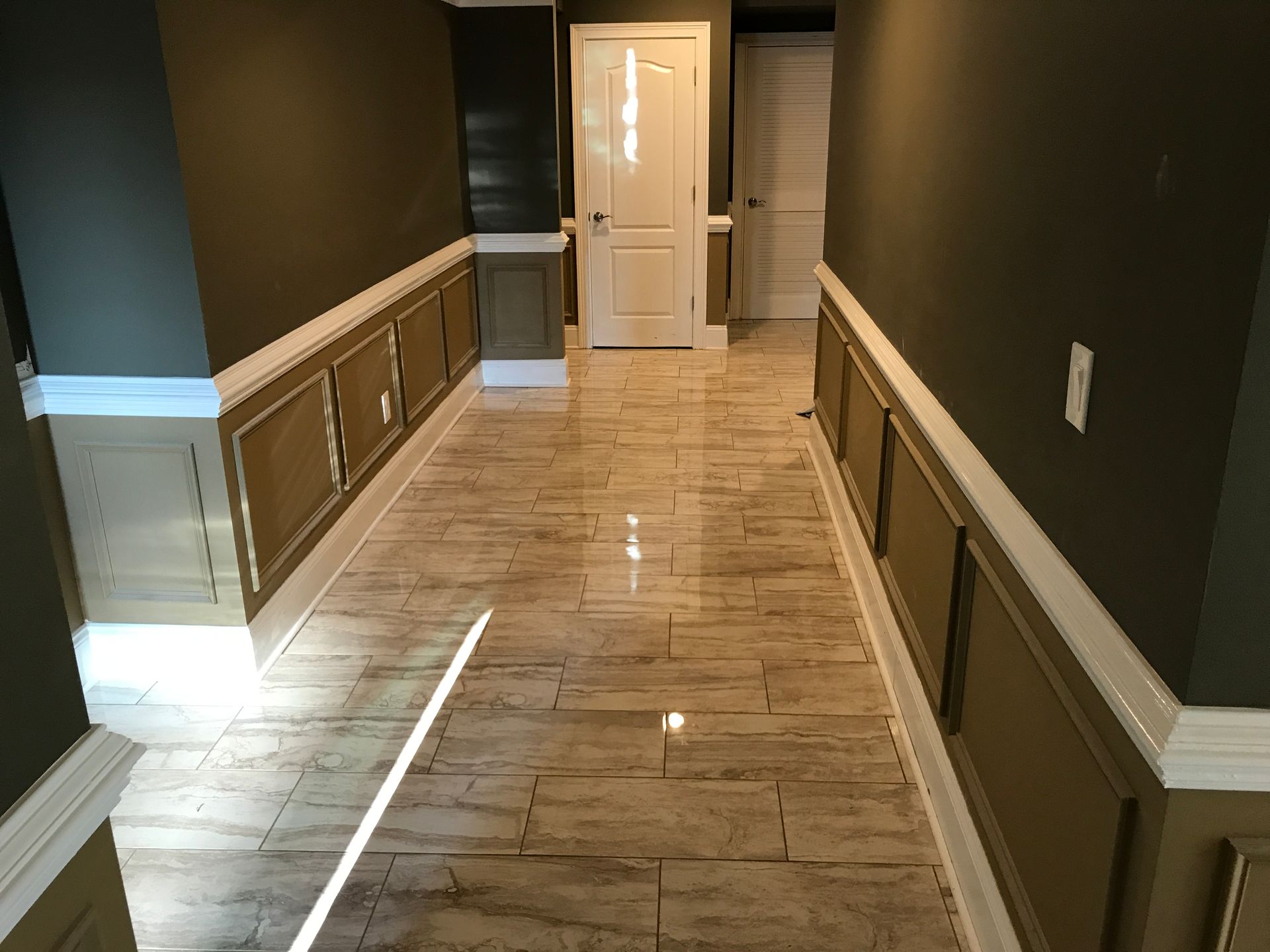 A hallway featuring polished wood-look tile flooring, dark olive walls with wainscoting, and two white doors at the end