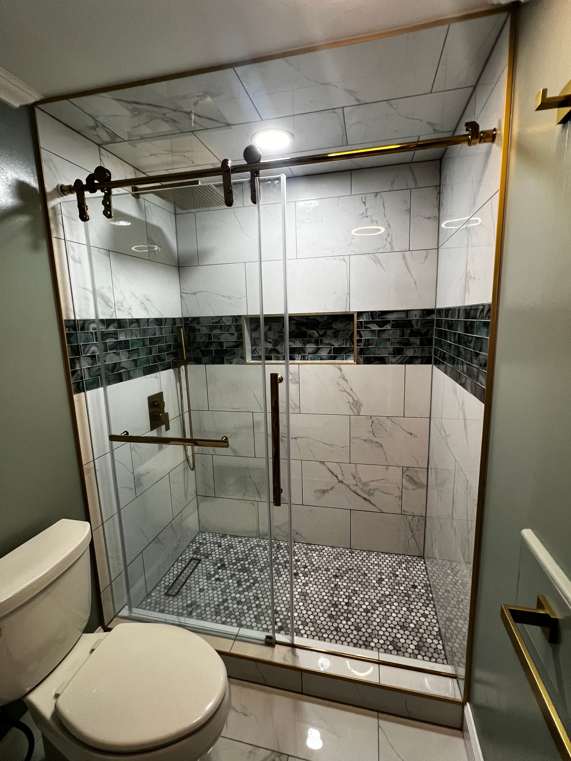 A walk-in shower with marble-style tiles, a decorative dark accent strip, glass sliding doors, and gold hardware