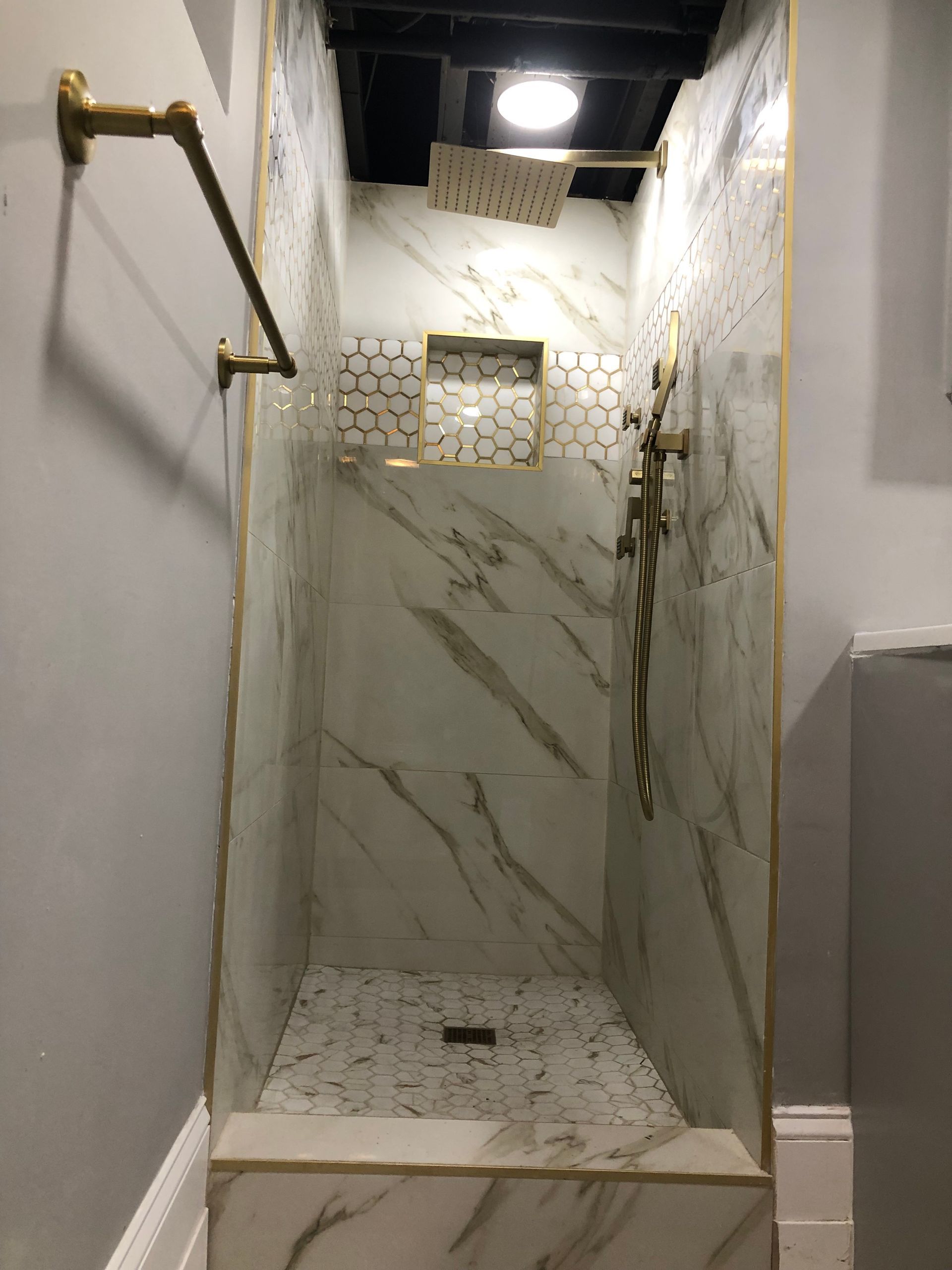 Small marble-tiled shower stall with glass shelf, wall grab bars, and overhead light