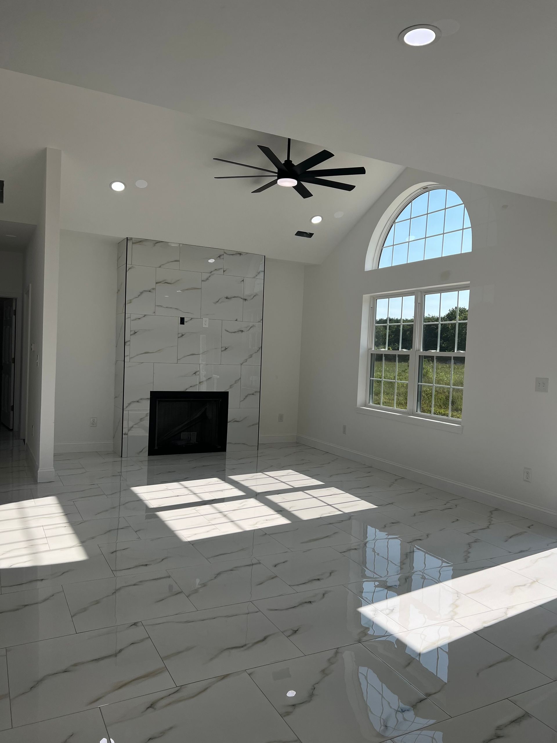 Bright empty living room with marble fireplace, arched window, ceiling fan, and sunlight on glossy floor
