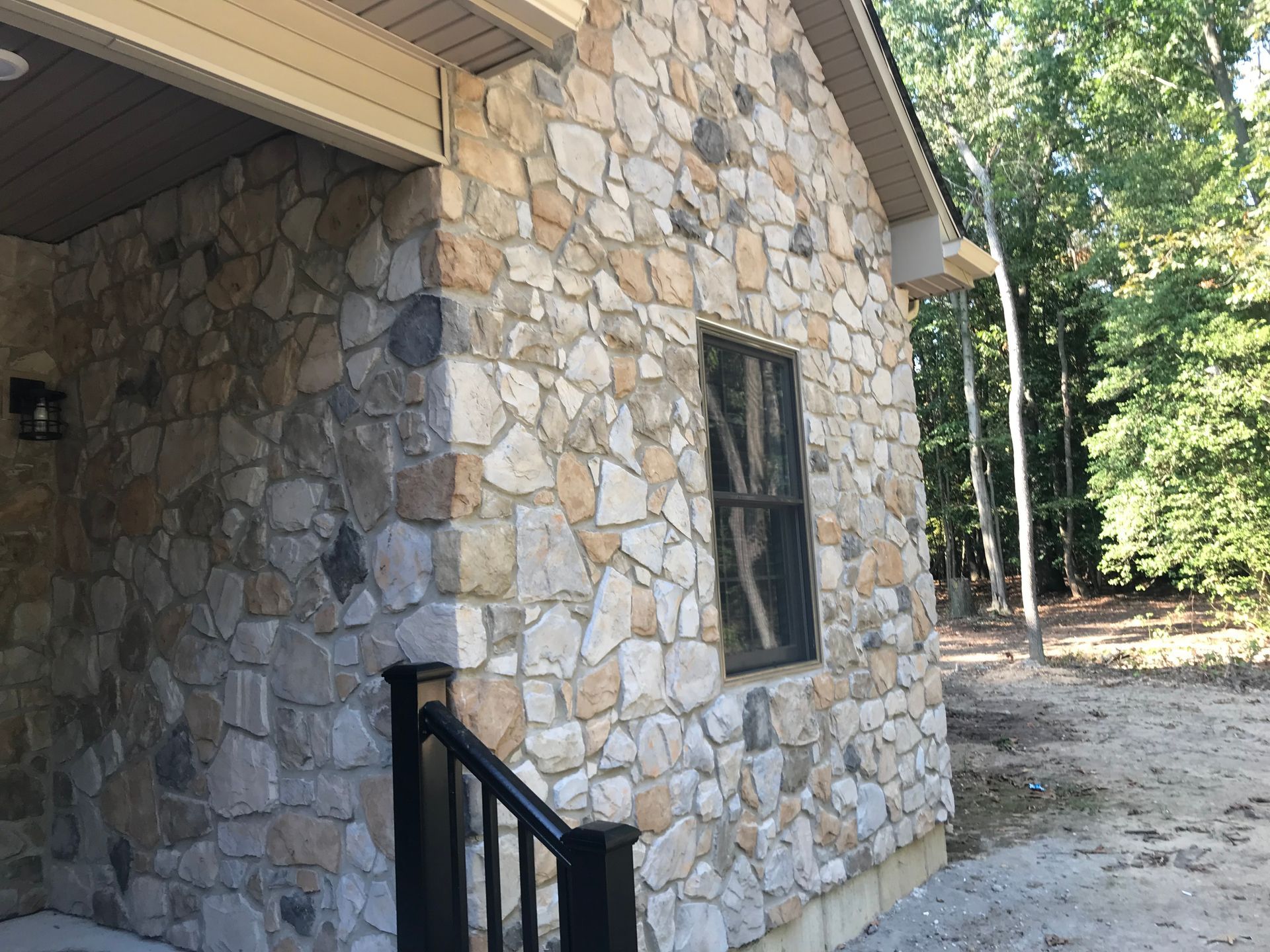 Stone house exterior with a black handrail, window, and wooded yard beside a gravel area