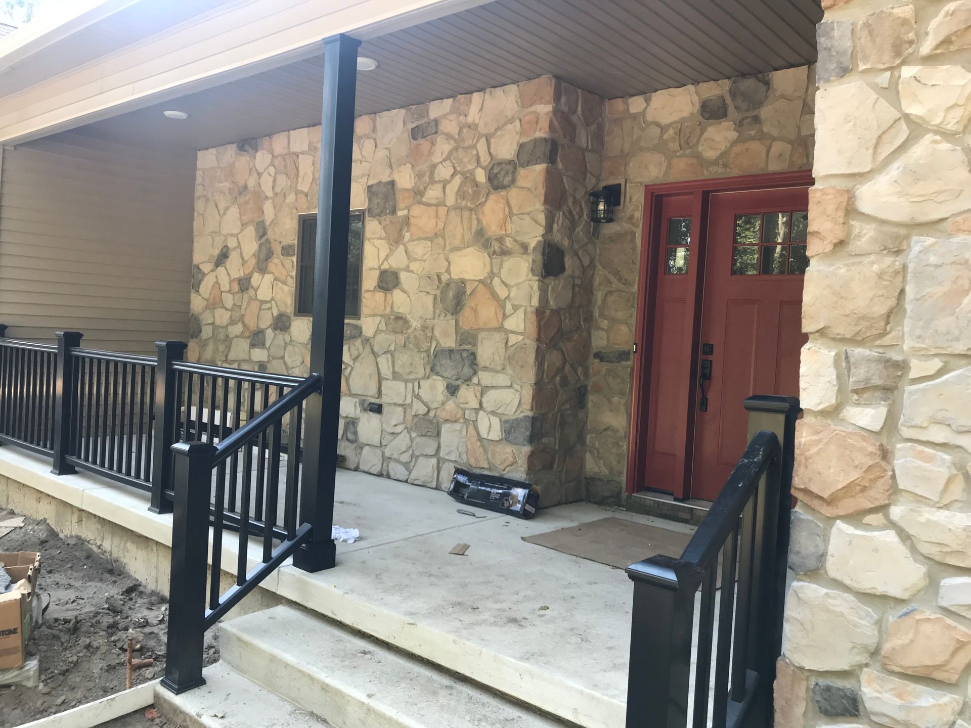 Front porch with stone walls, red door, black railings, and concrete steps under a covered roof