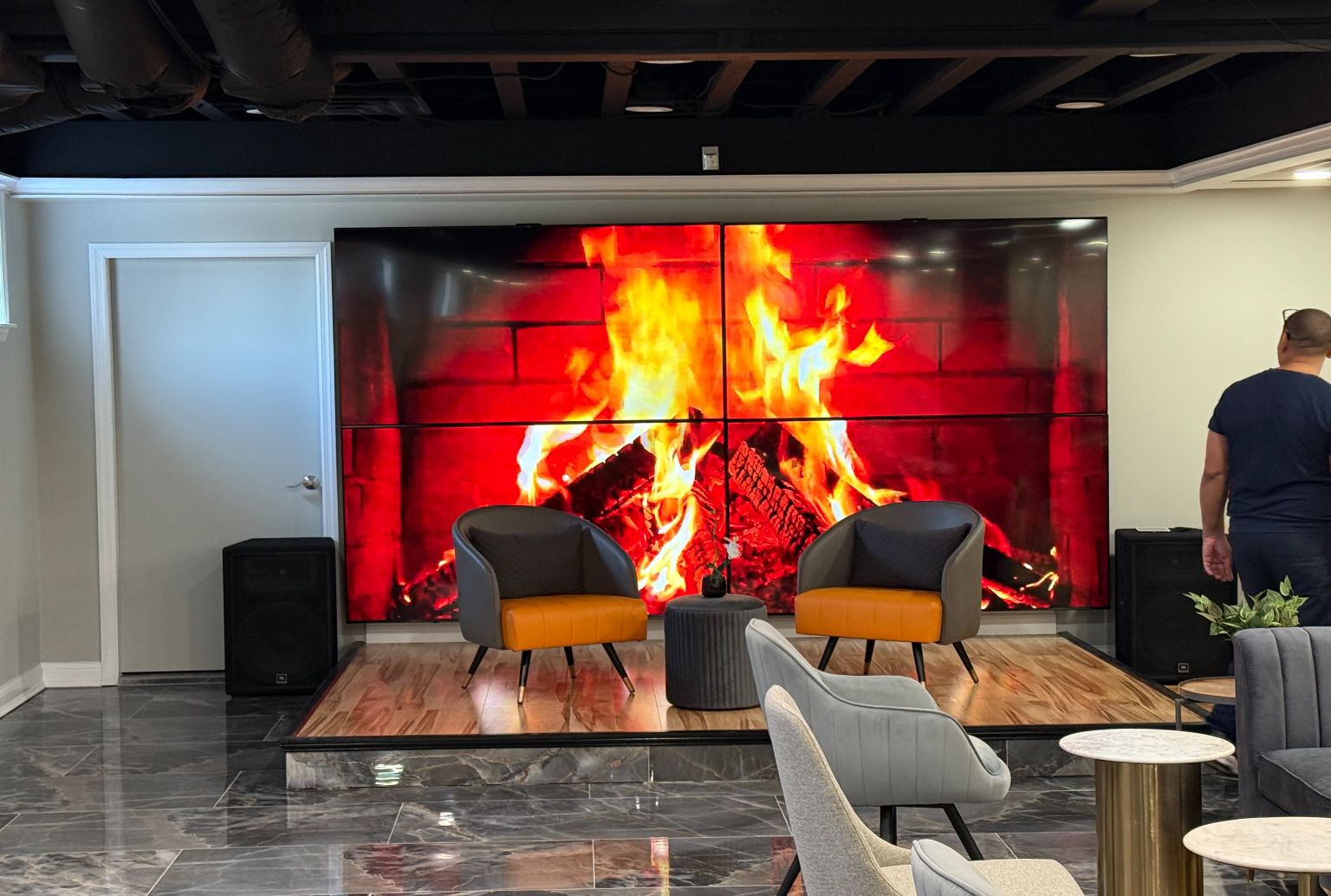 Two chairs and a small ottoman on a raised wooden platform in front of a wall-sized digital screen displaying a fireplace
