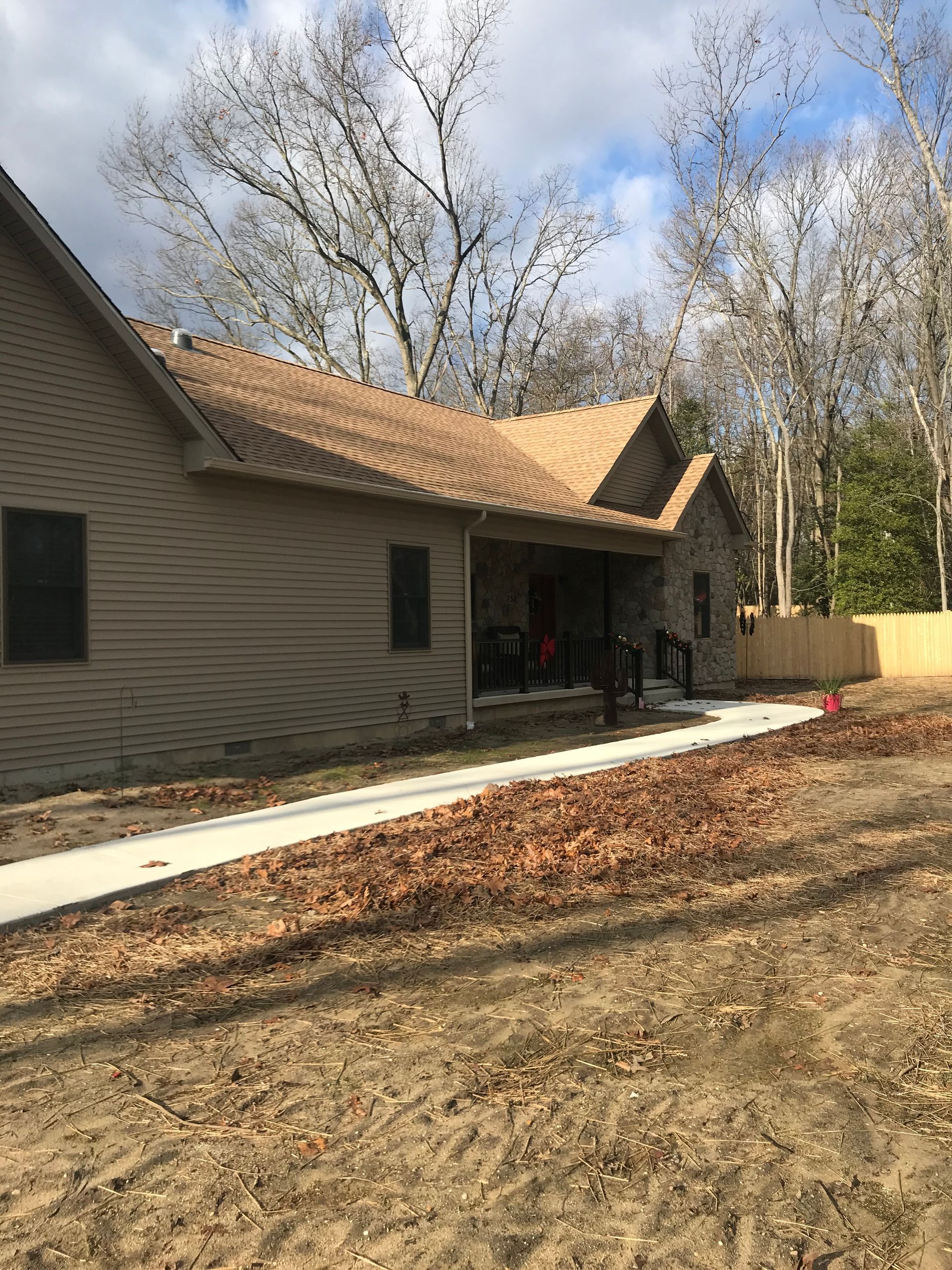 Suburban house with tan siding and roof, front porch, and bare trees in a yard with mulch and a sidewalk