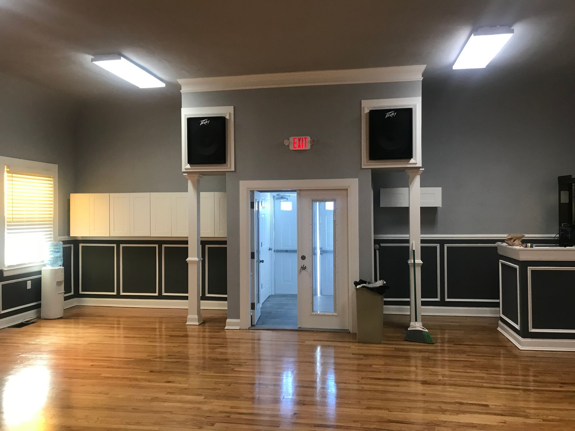 Empty event hall with wooden floor, gray walls, double doors, and large wall-mounted speakers