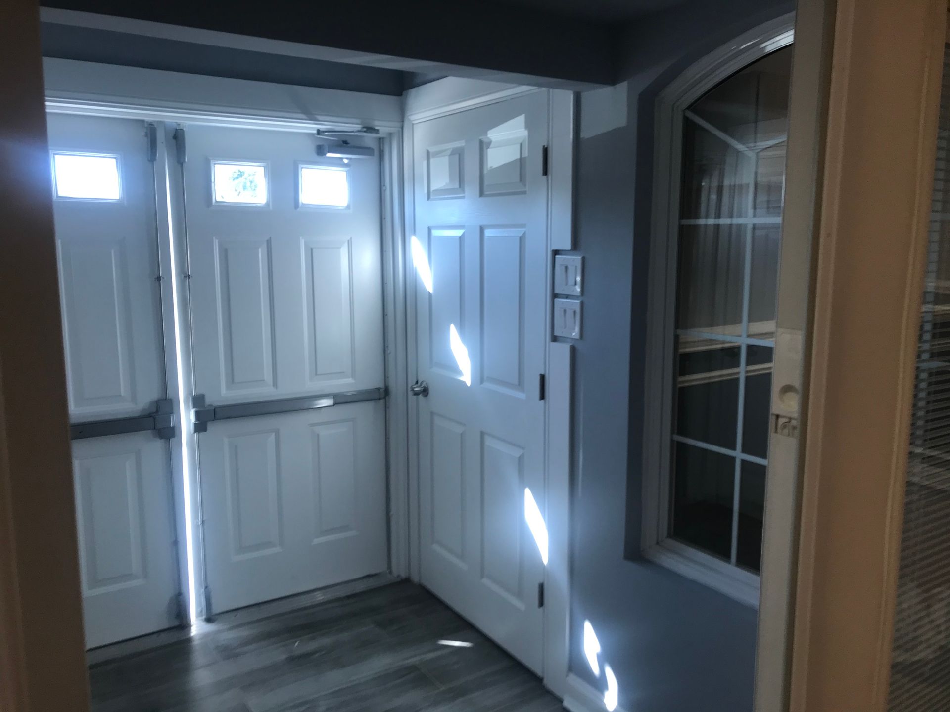 Front entryway with white double doors, side window, and gray wood floor, lit by sunlight