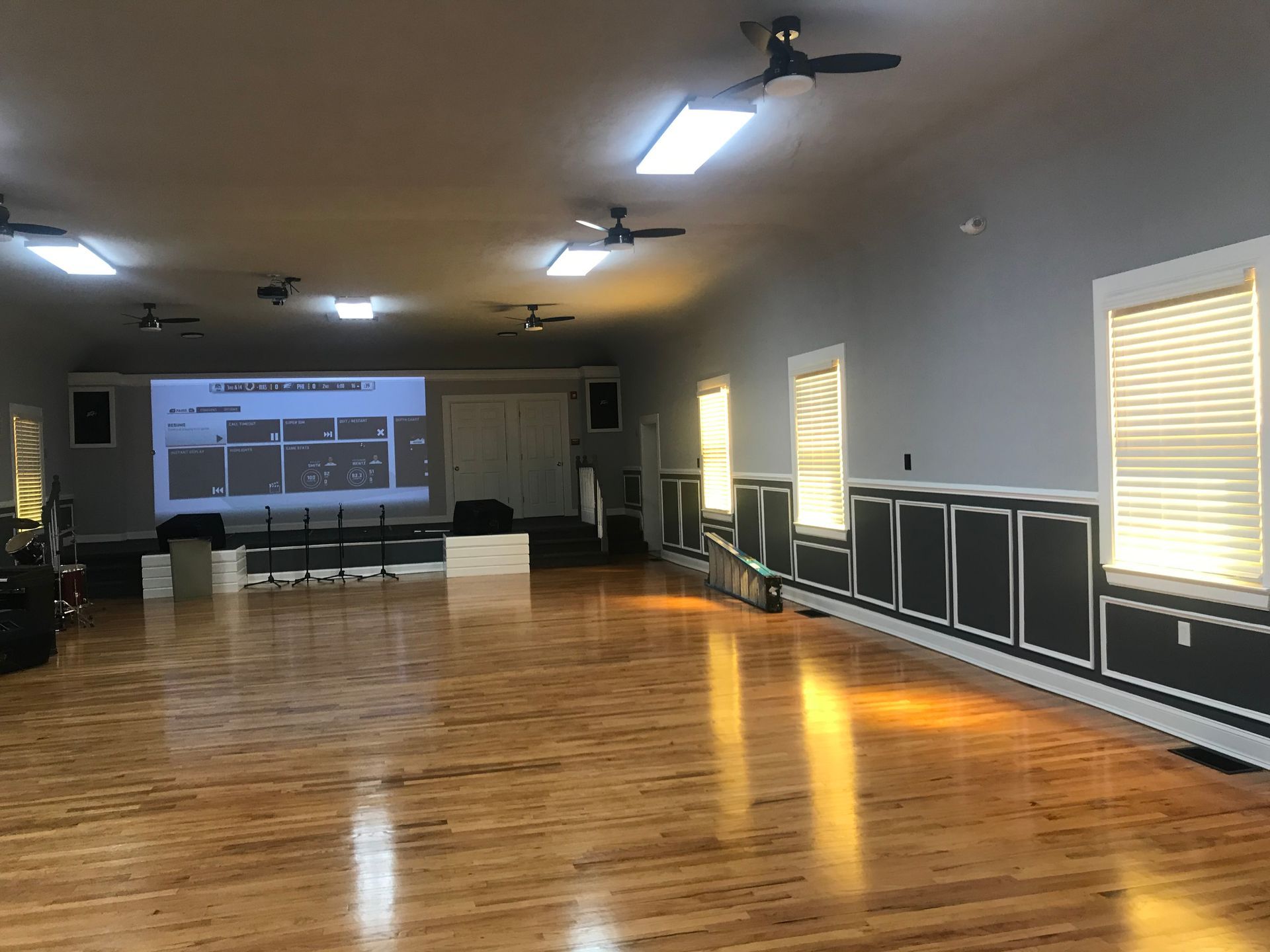 Empty gym with polished wooden floor, wall mirrors, windows, and a projector screen at the far end