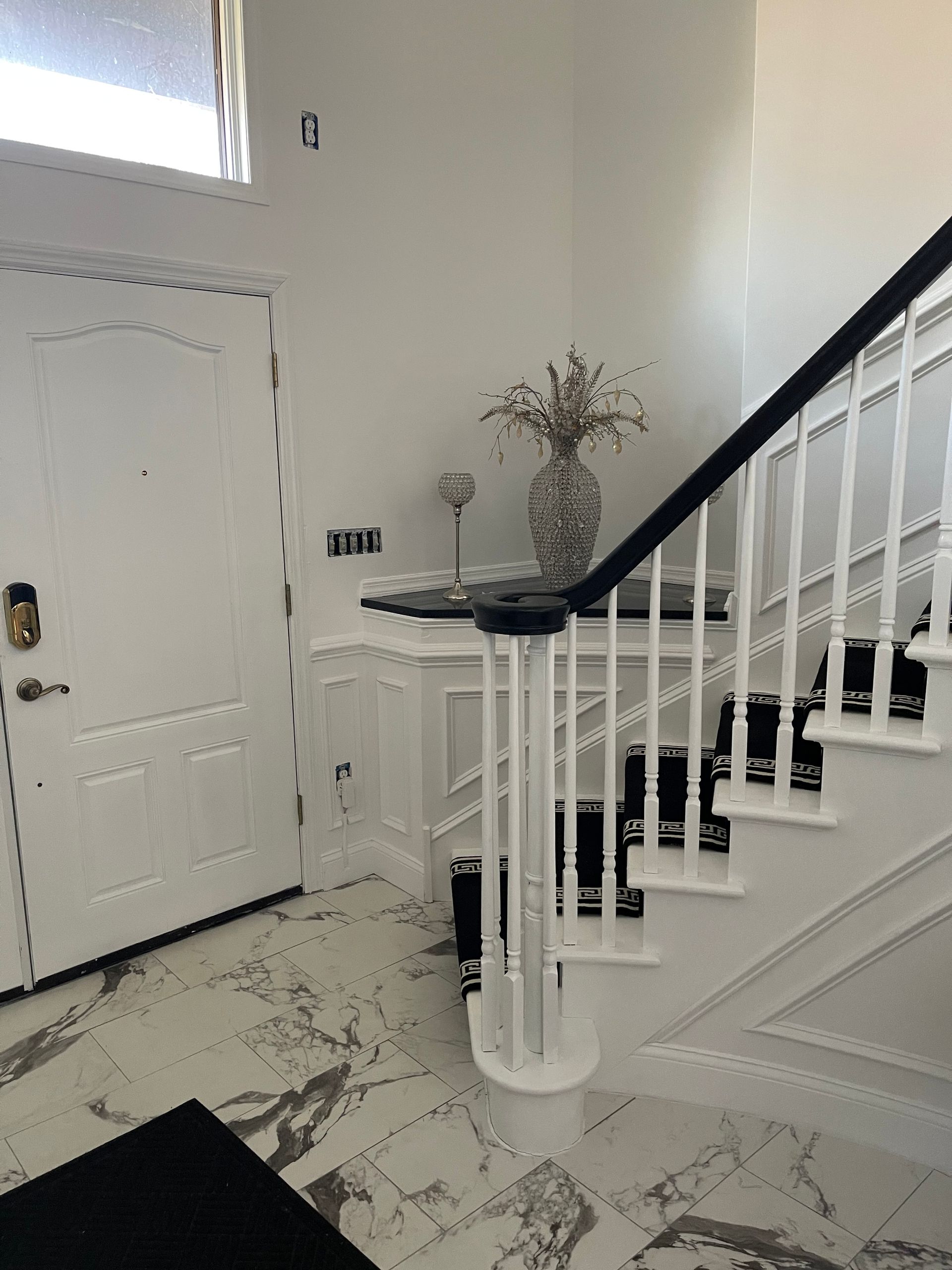 Bright entryway with white staircase, front door, marble floor, and a vase on a small table
