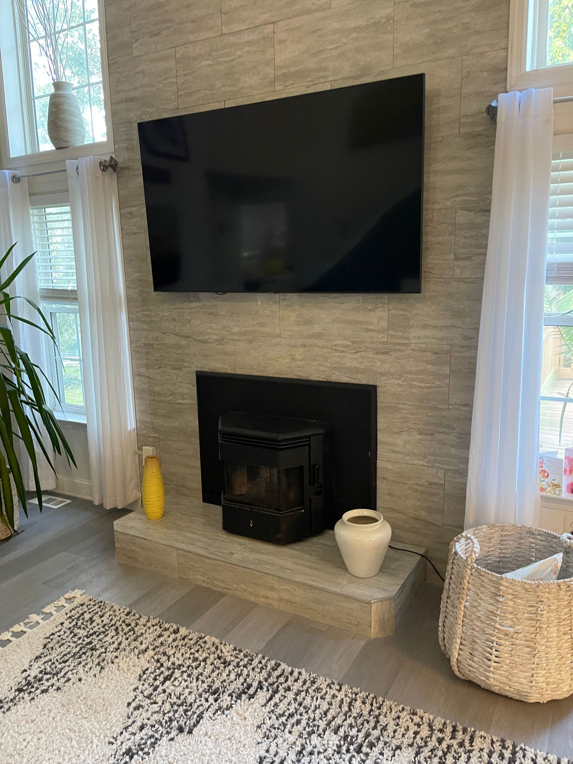 Modern living room with wall-mounted TV above a black fireplace, flanked by tall windows and wicker baskets