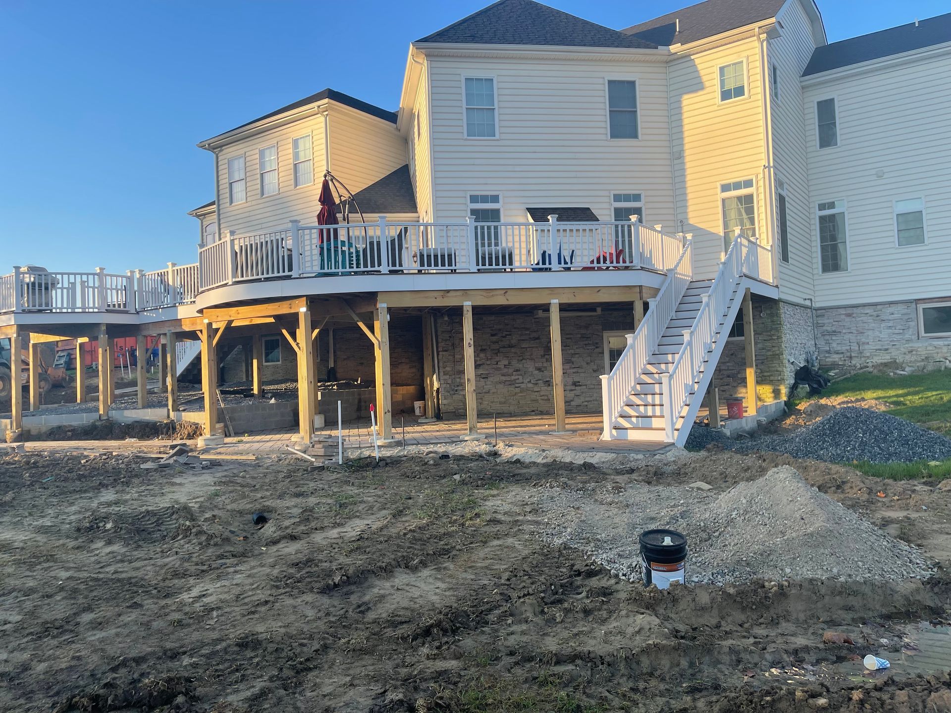 A large beige house with a wrap-around wooden deck and white railings under a clear blue sky, with a dirt yard below