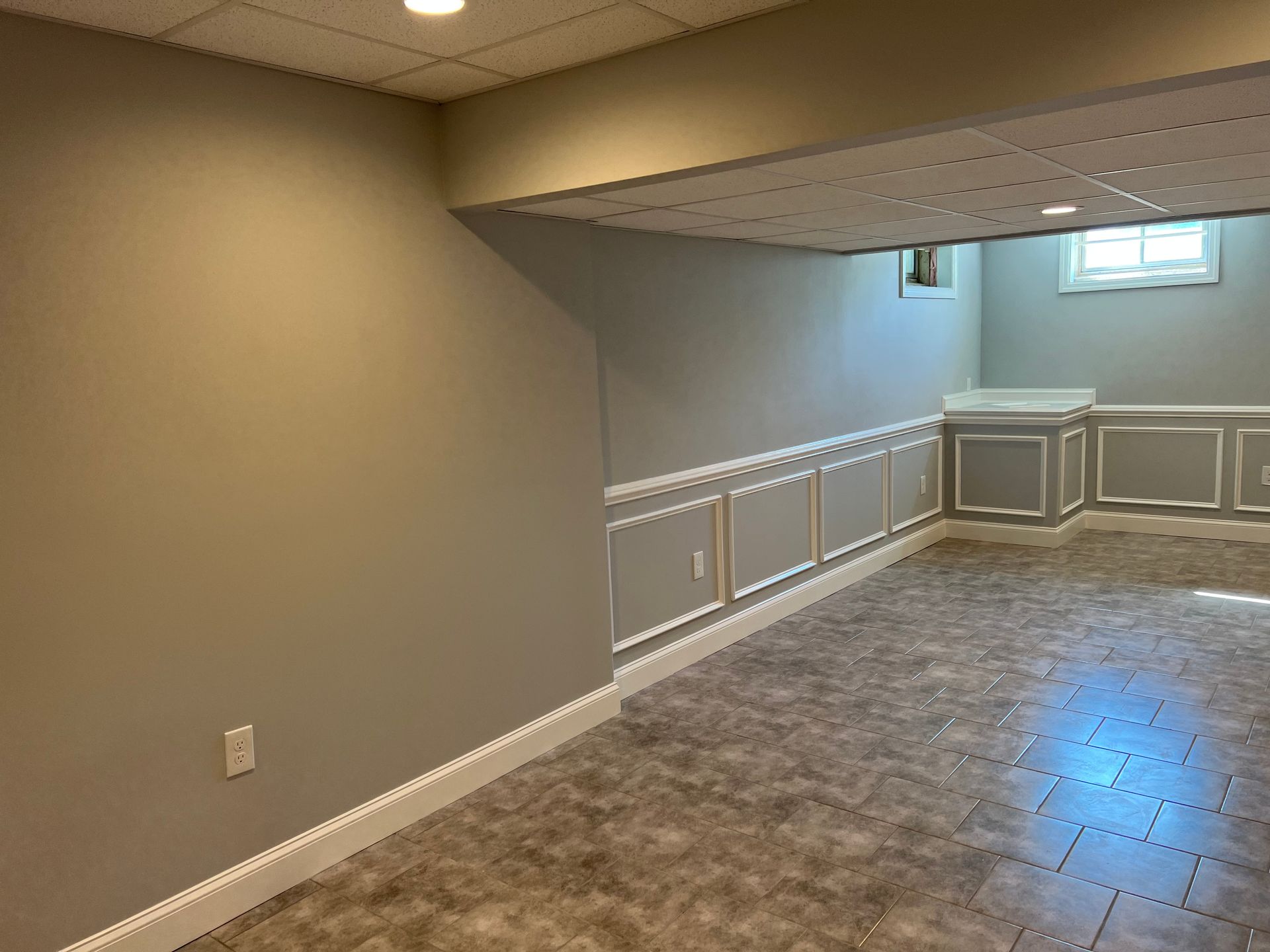 Empty carpeted basement room with gray walls, tiled floor, and a low ceiling with windowed railing