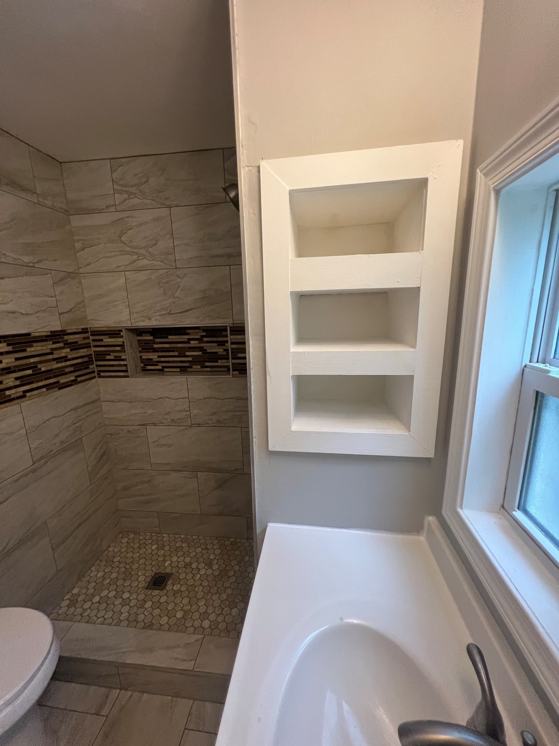 Small bathroom with shower, tub, window, and white wall shelves above the tub